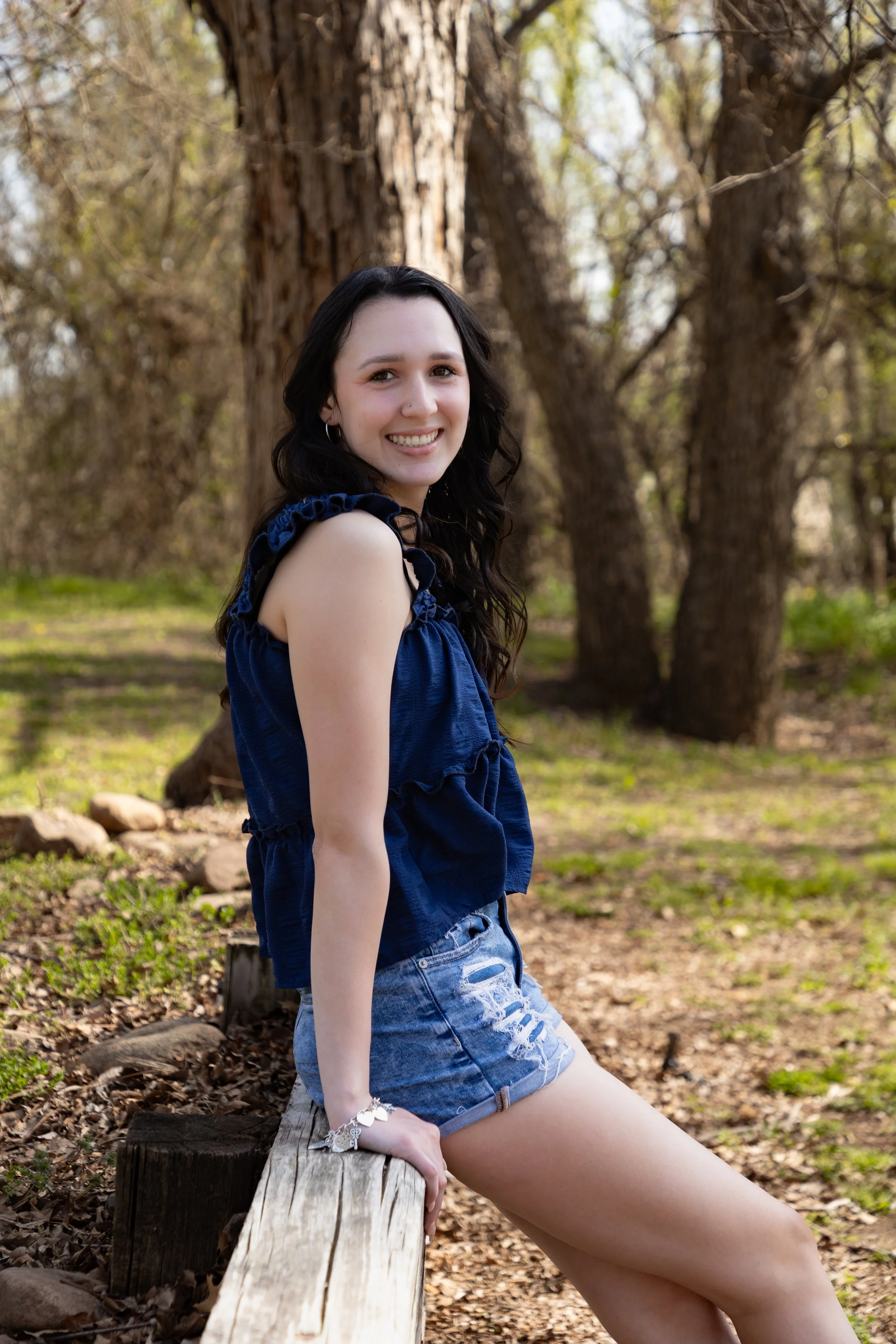 A young woman with dark curly hair, smiling, outdoors in a wooded area, sitting on a tree log, wearing a blue sleeveless top and distressed denim shorts.