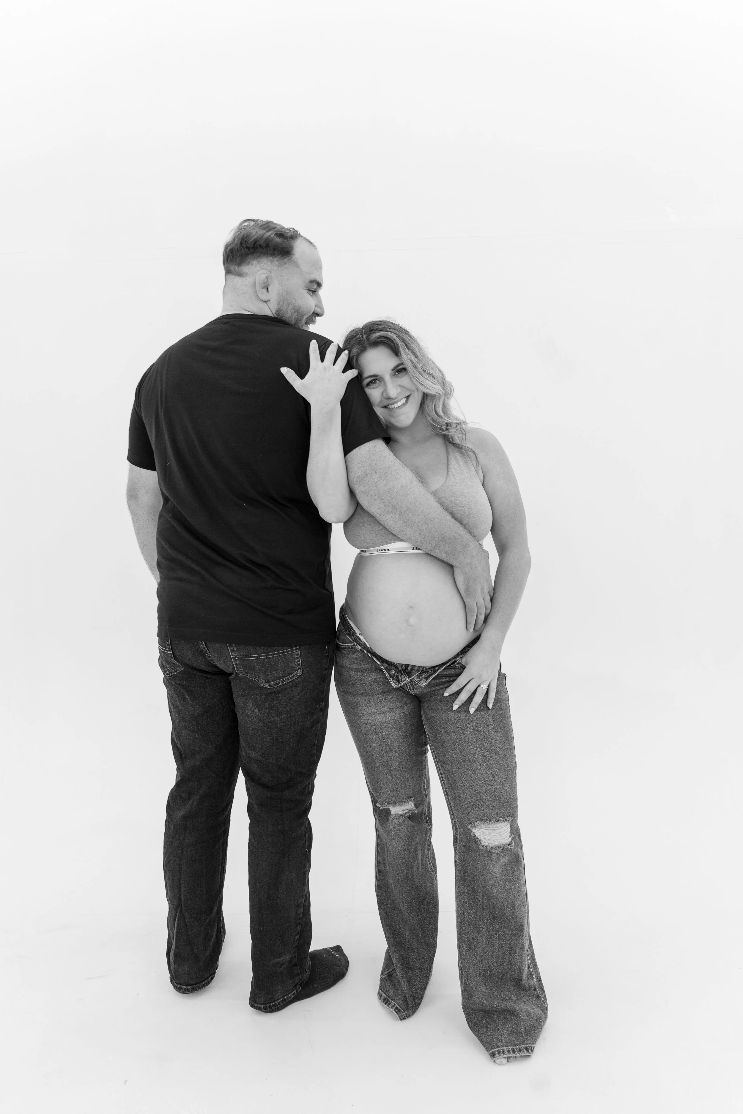 A black-and-white photo of a man and woman smiling and embracing against a plain white wall. The woman has long curly hair and is wearing a crop top and ripped jeans, while the man has short hair, a beard, and is wearing a t-shirt and jeans.