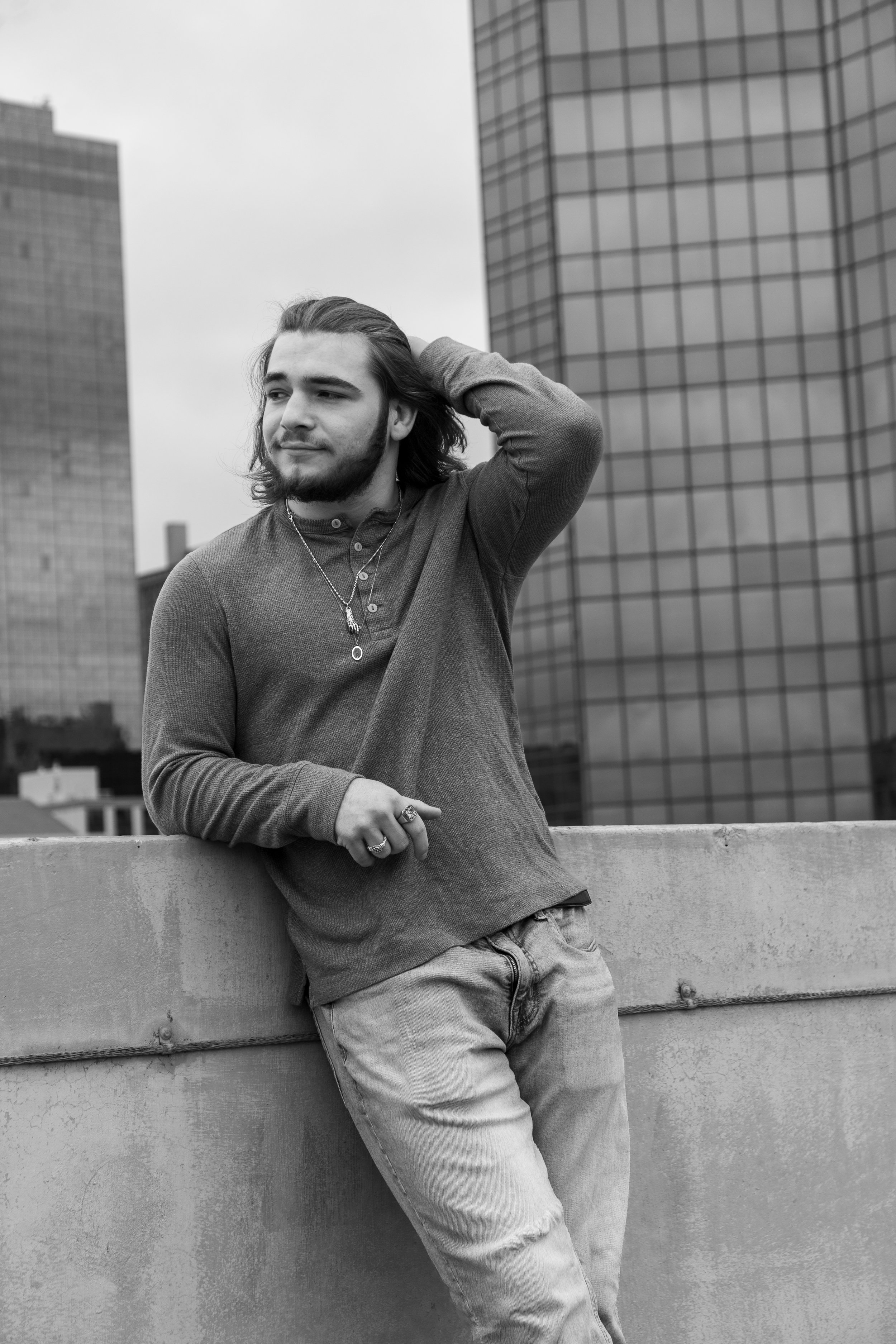 A young man with long hair and a beard standing on a rooftop, leaning against a concrete barrier with skyscrapers in the background, dressed in a long-sleeve shirt and light jeans, looking thoughtfully to the side.