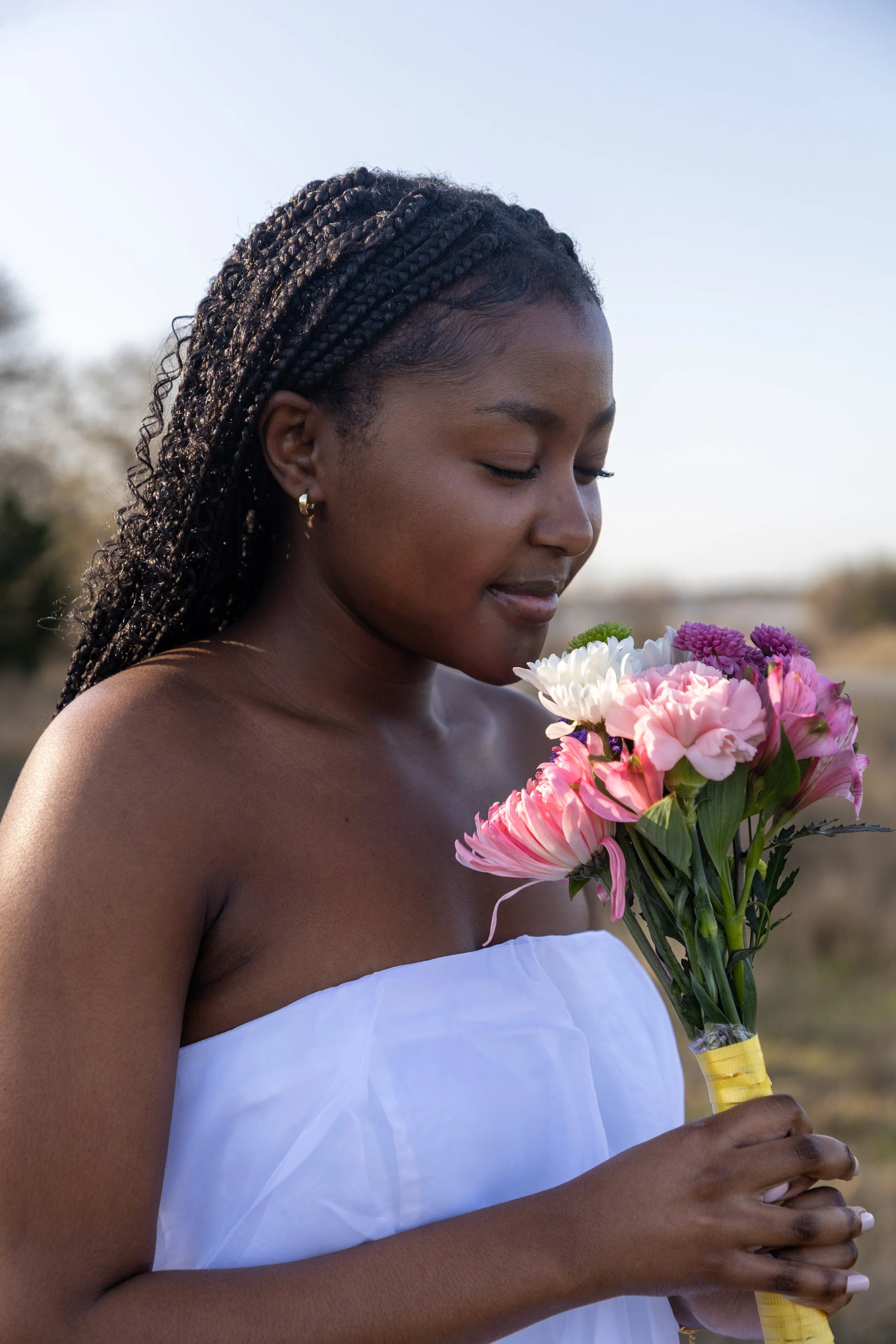 A woman with braids and earrings, wearing a white strapless dress, holding a bouquet of pink, white, and purple flowers outdoors.