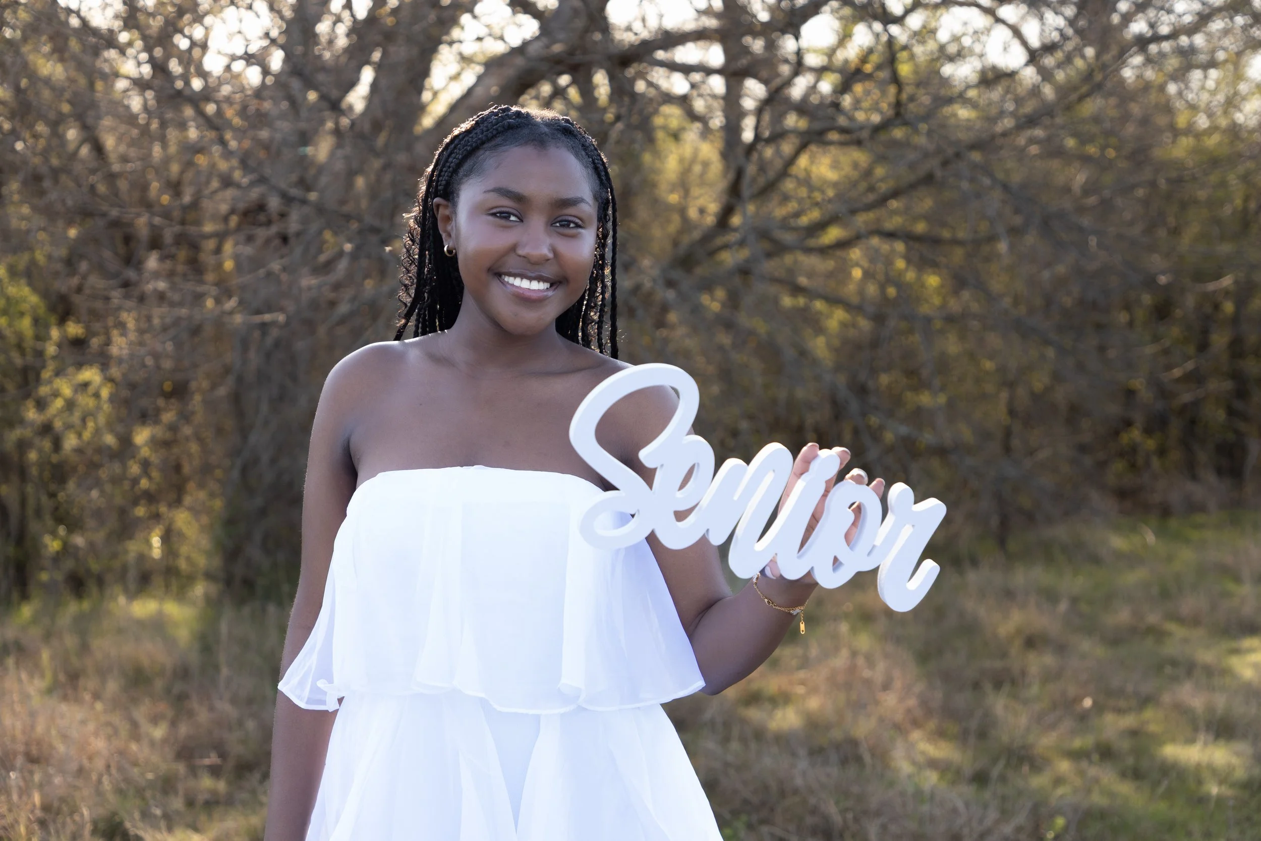 Young girl smiling outdoors holding a white sign that says 'Senior' in a nature setting with trees in the background.