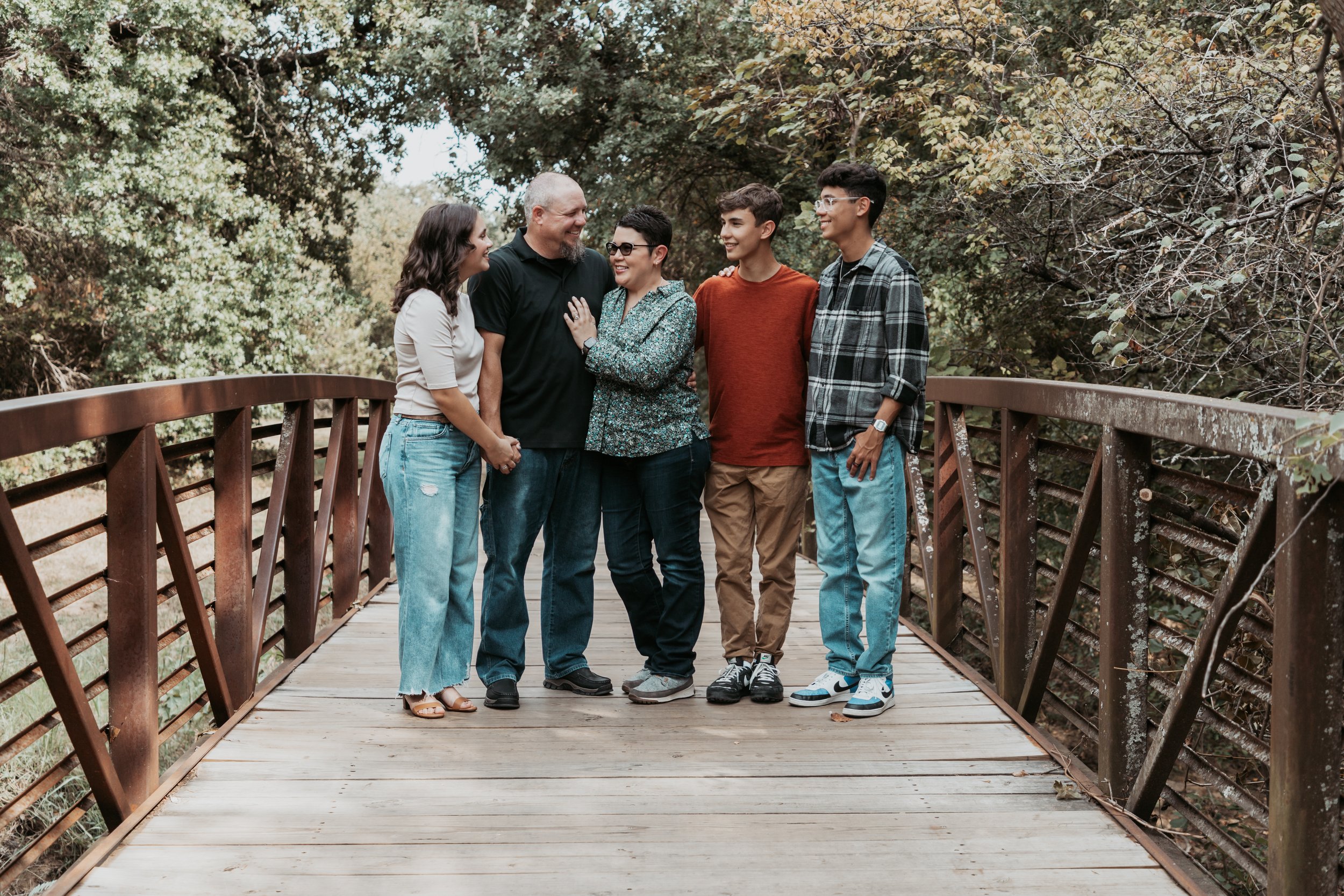 Group of six people standing on a wooden bridge outdoors, smiling and talking in a natural setting with trees in the background.