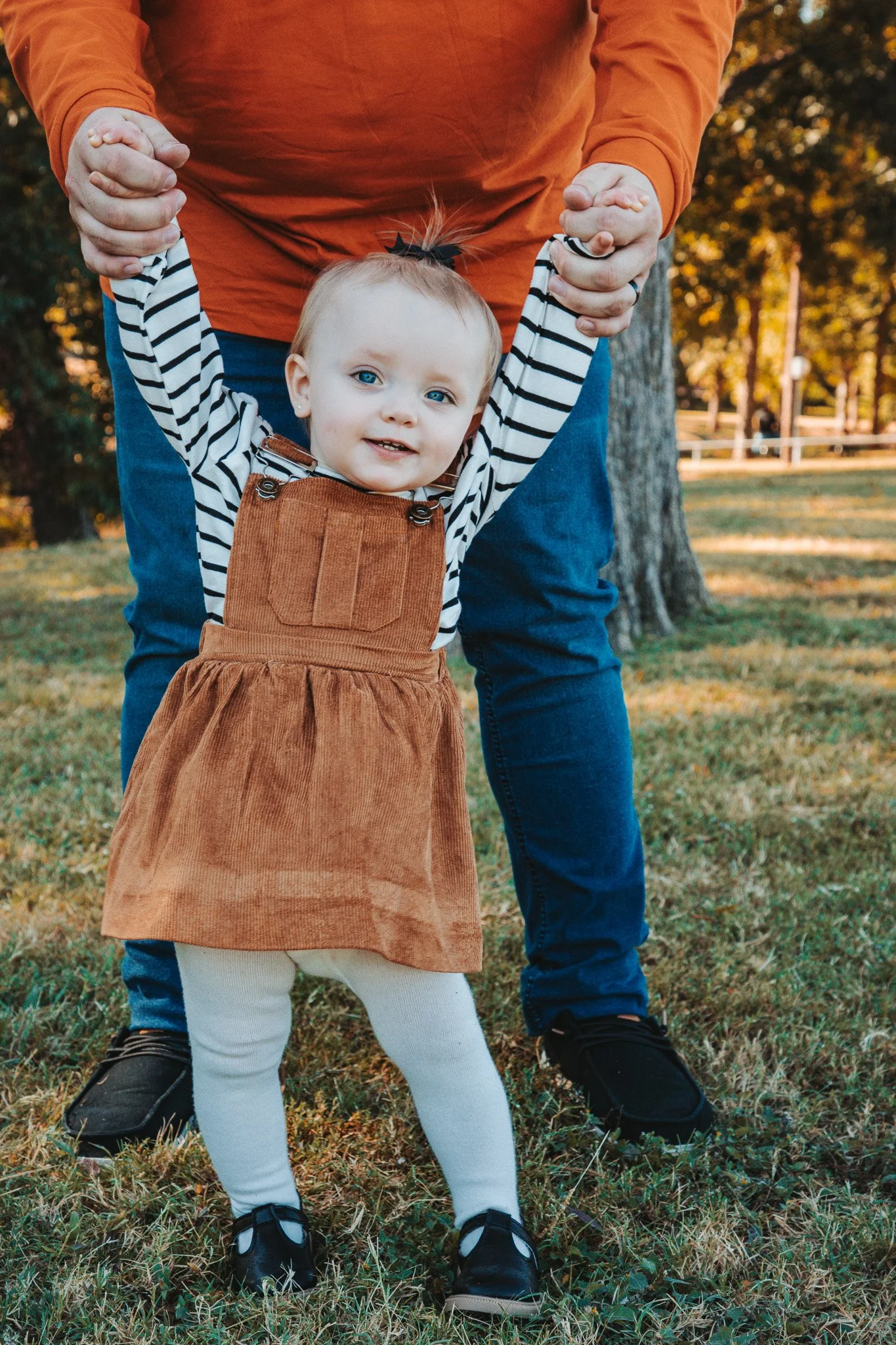 A young girl with blonde hair, wearing a brown corduroy dress over a black and white striped long-sleeve shirt, standing outside on grass while holding onto an adult's hands, with trees and a fence in the background.