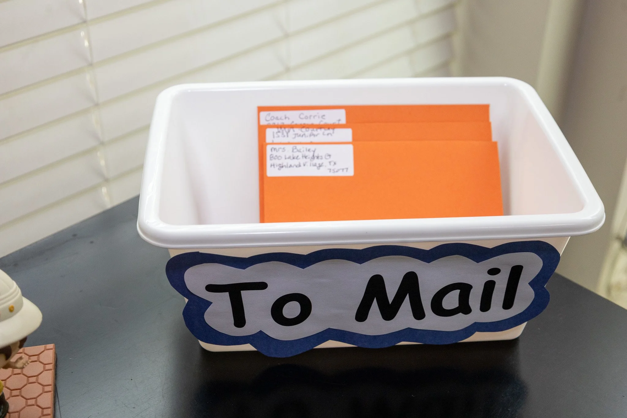 A white plastic mailbox with a blue and gray 'To Mail' sign on the front, containing orange envelopes with handwritten addresses.