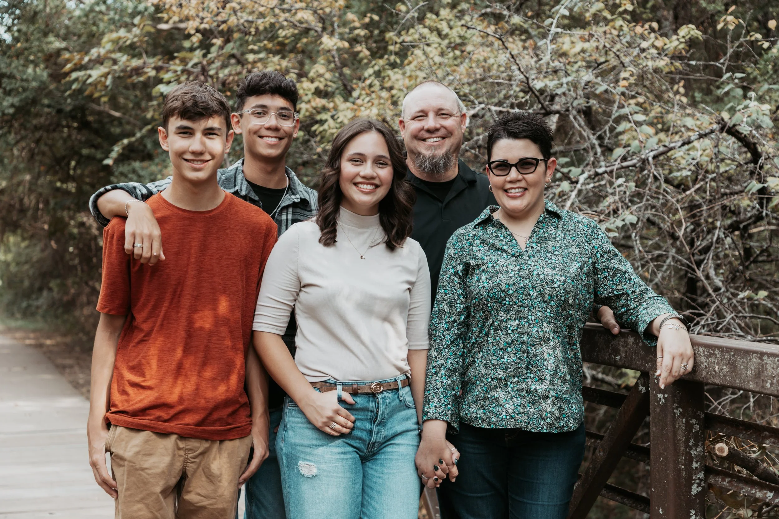 A family of six standing outdoors on a wooden bridge, with trees and foliage in the background, smiling at the camera.