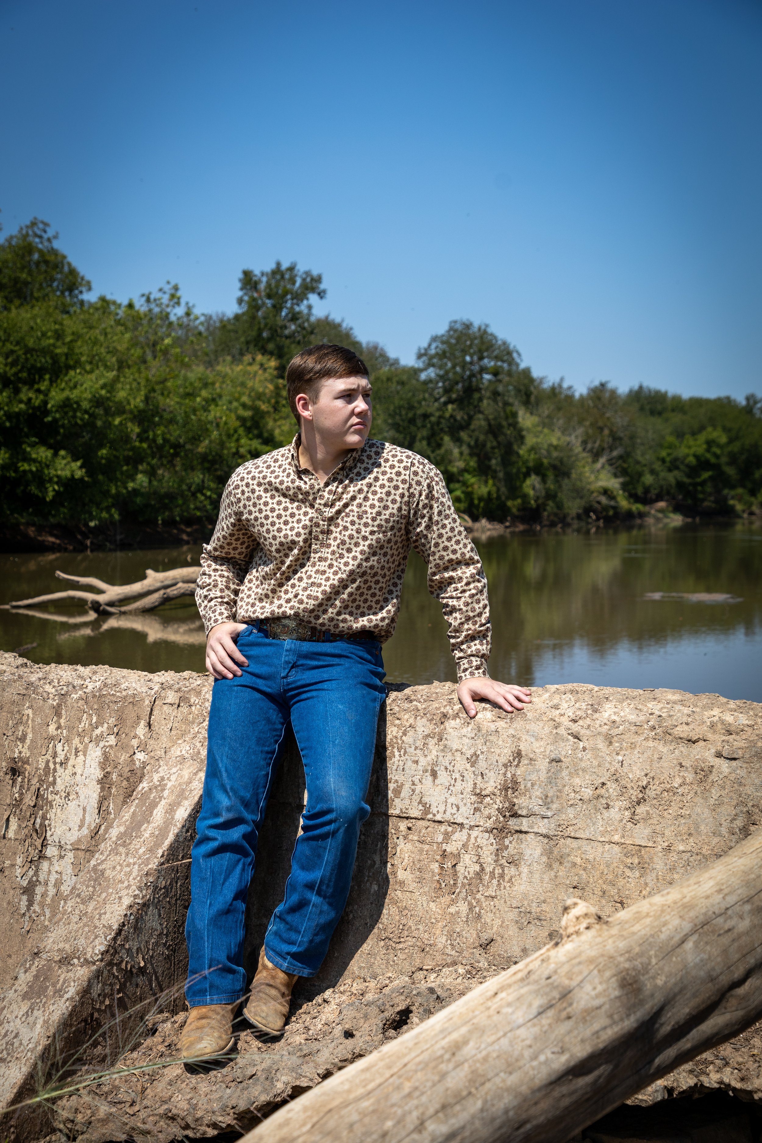A young man in a patterned shirt and jeans standing on a large rock near a river, with trees and a clear blue sky in the background.