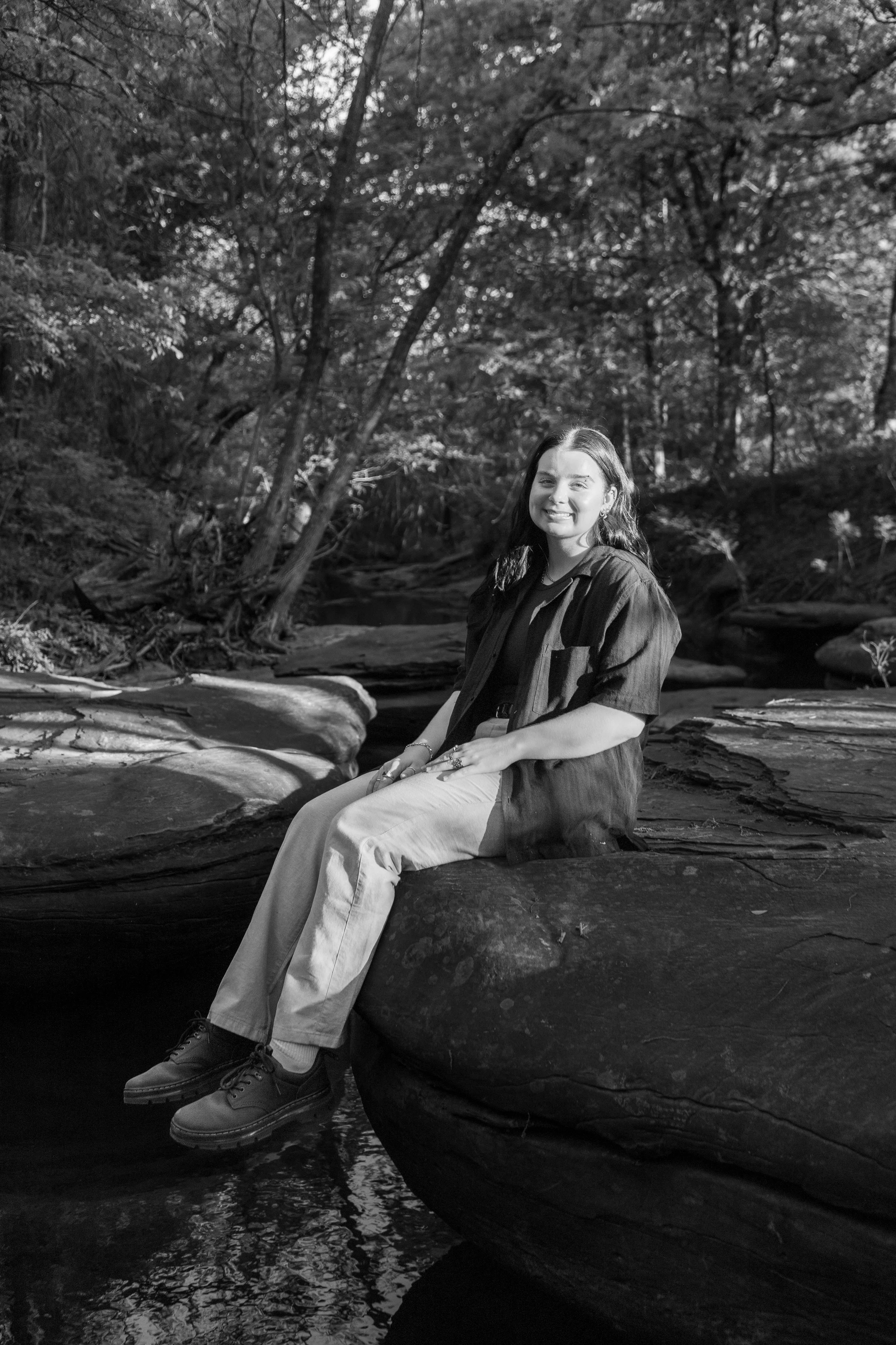 A young woman sitting on a rock by a creek in a wooded area.
