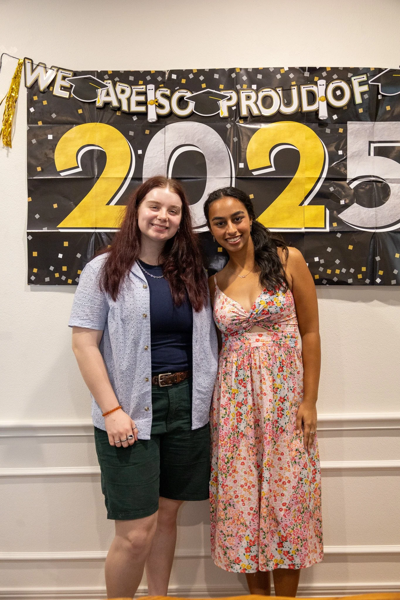 Two women standing together at a graduation celebration, in front of a black and gold decorated banner that says, "We Are So Proud Of 2023."