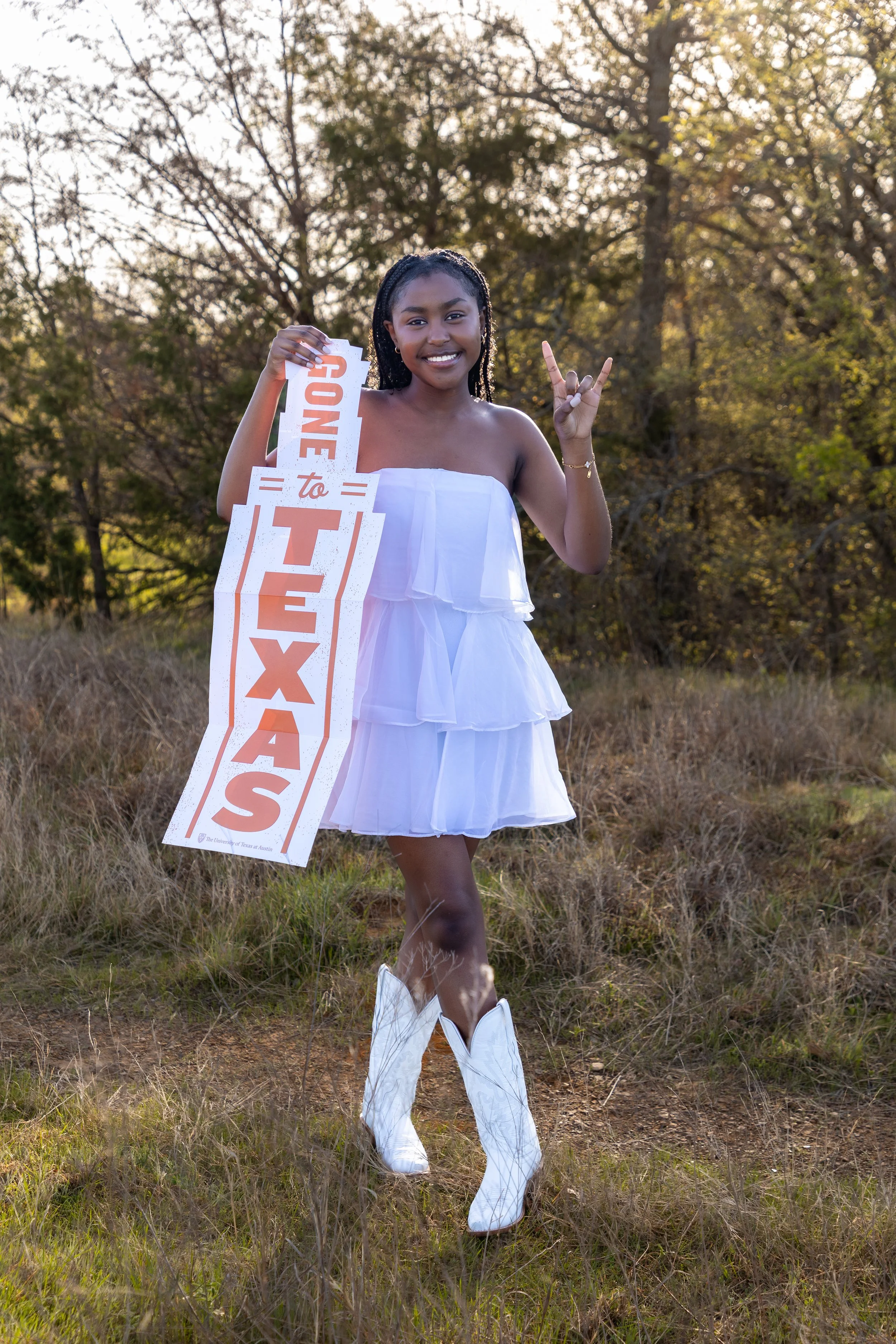 Young woman standing outdoors in a grassy area holding a sign supporting a cause, making a rock and roll hand gesture, wearing a white dress and cowboy boots, with trees in the background.