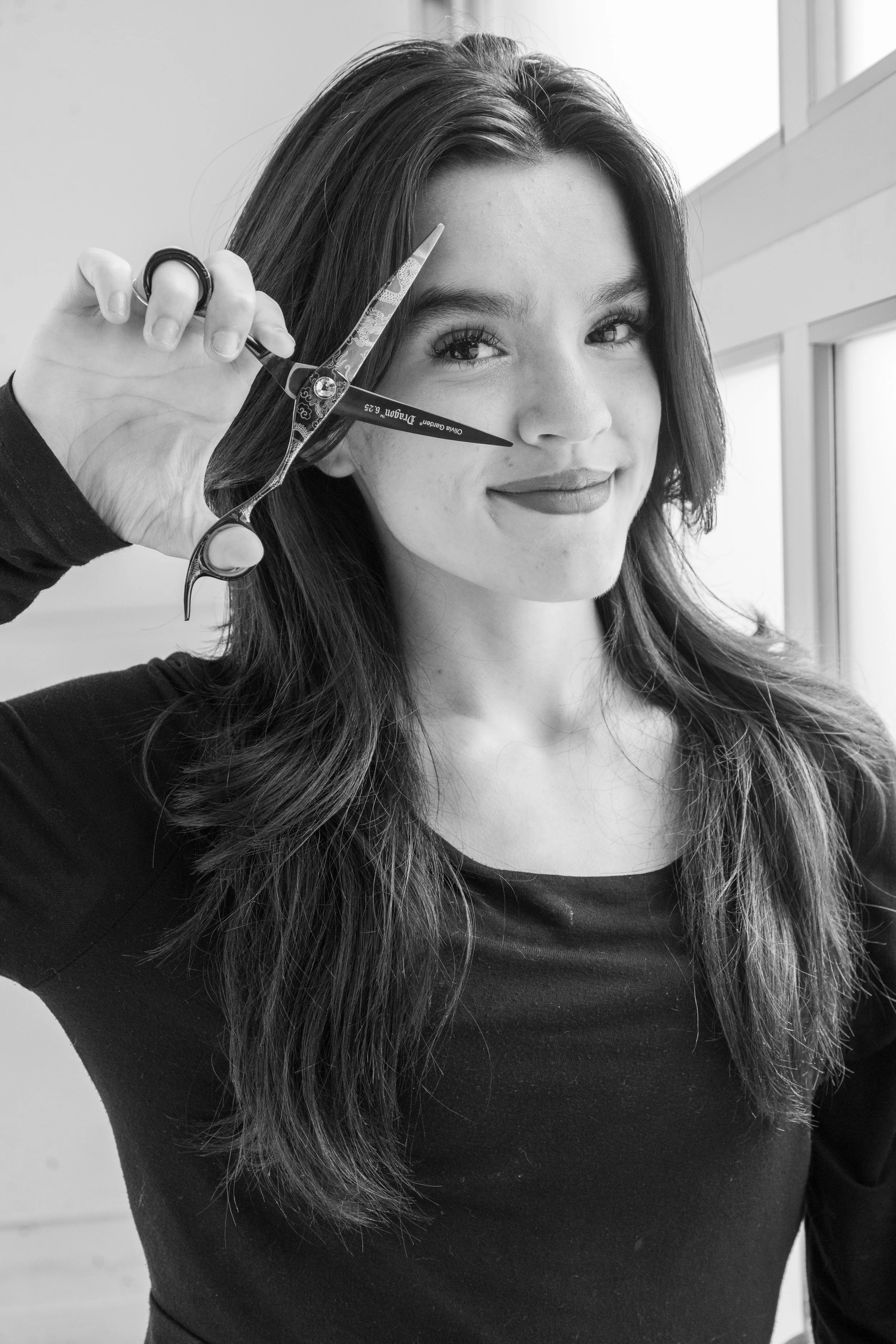 Black and white photo of a young woman with long wavy hair, smiling at the camera, holding a pair of scissors near her face, with a window in the background.
