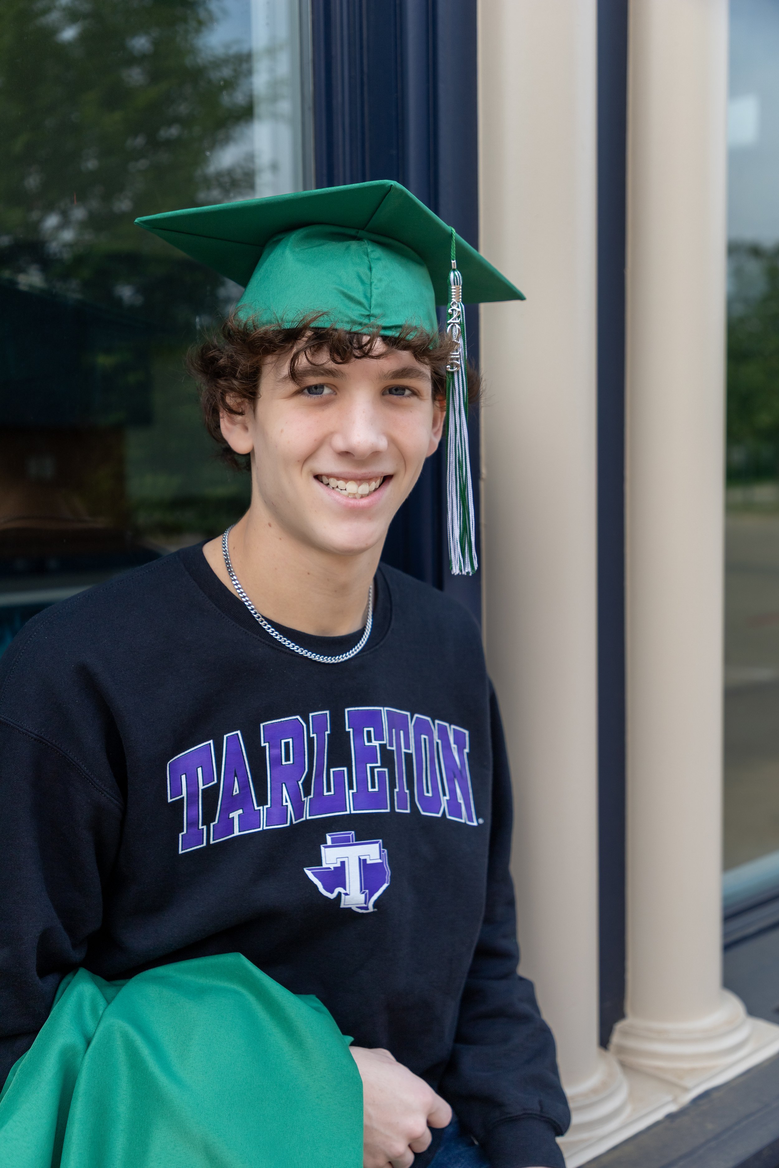 A young man smiling in a black Tarlton sweatshirt wearing a green graduation cap, holding a green bag, standing outside near a building.