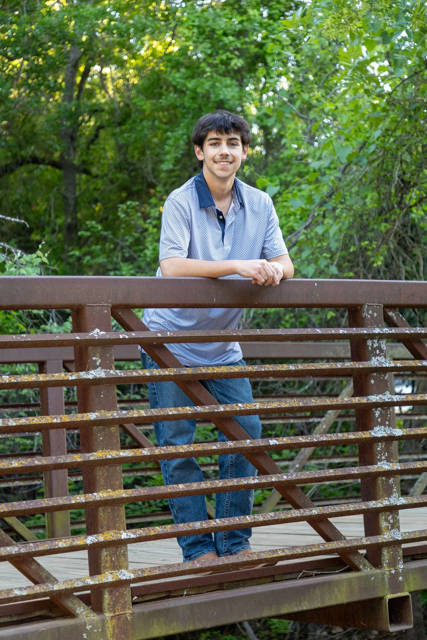 A young man with dark hair smiling while leaning on a rusty metal bridge railing in a wooded area.