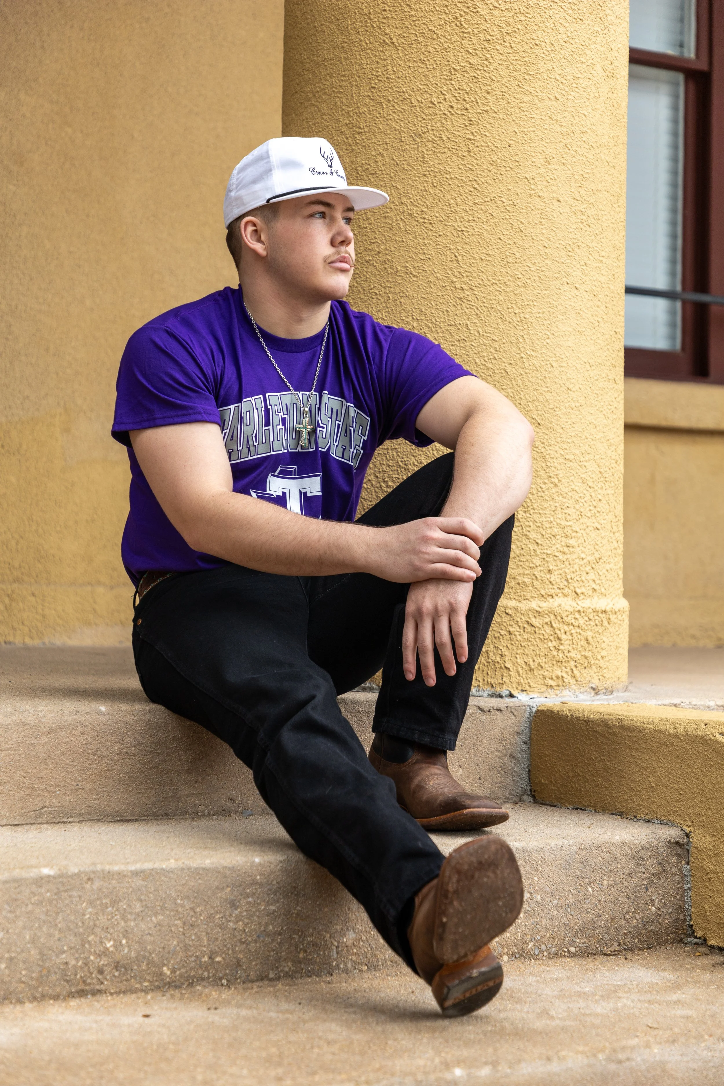 Young man sitting on outdoor steps, wearing a purple TCU shirt, white cap, black jeans, brown boots, and a silver chain, leaning against a yellow textured wall with a window nearby.