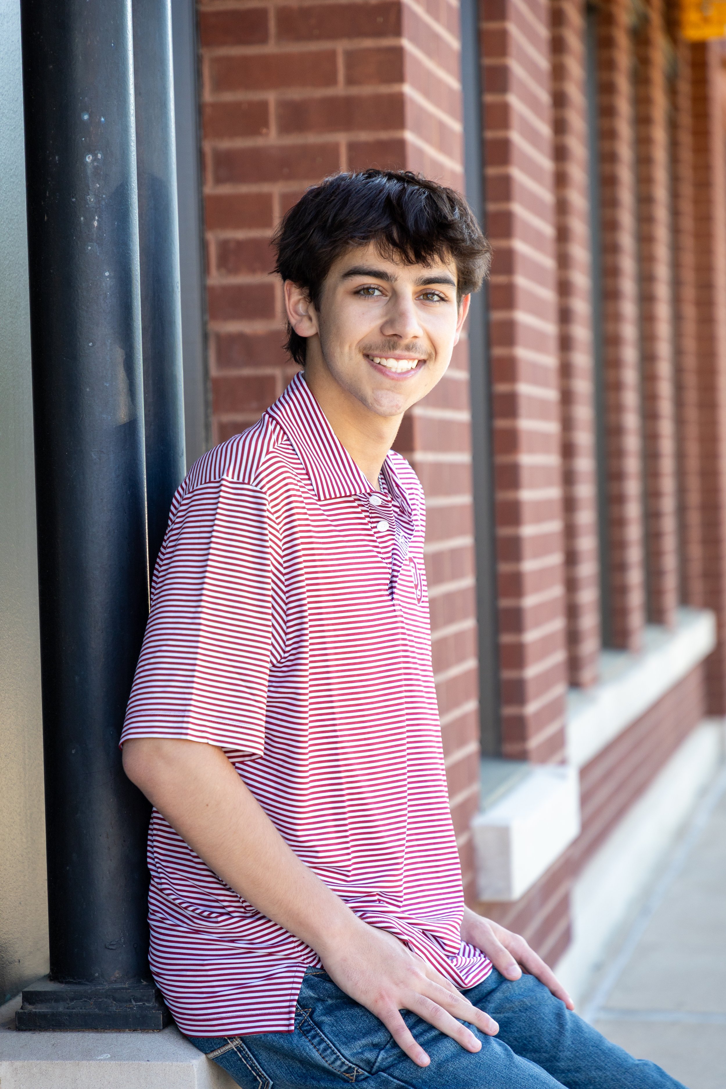 A young man with dark hair and a smile, wearing a red and white striped shirt, leaning against a black pole outside a brick building.