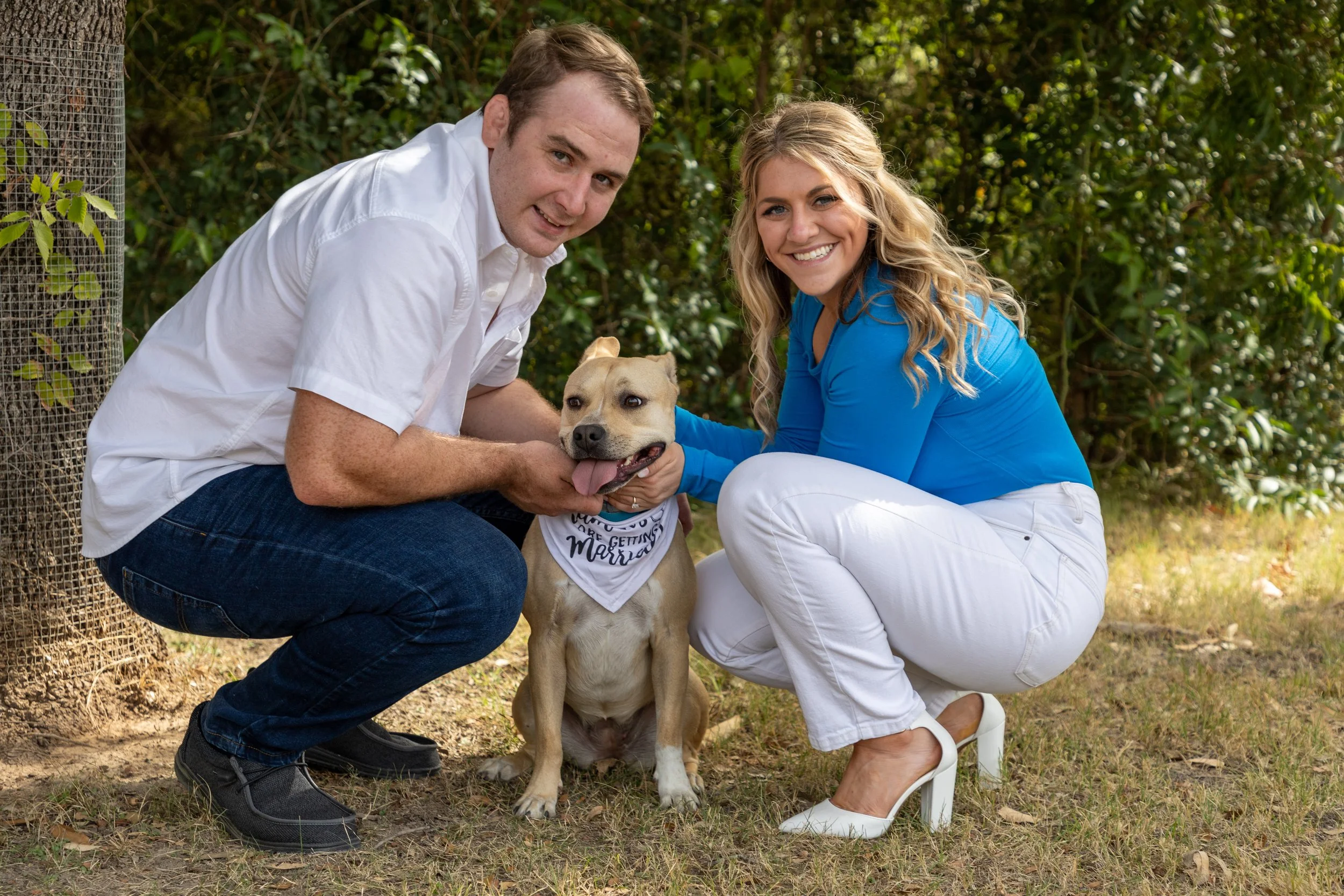 A man and woman smiling and holding a happy tan and white dog with a bandana on its neck outdoors on grass, with green bushes in the background.