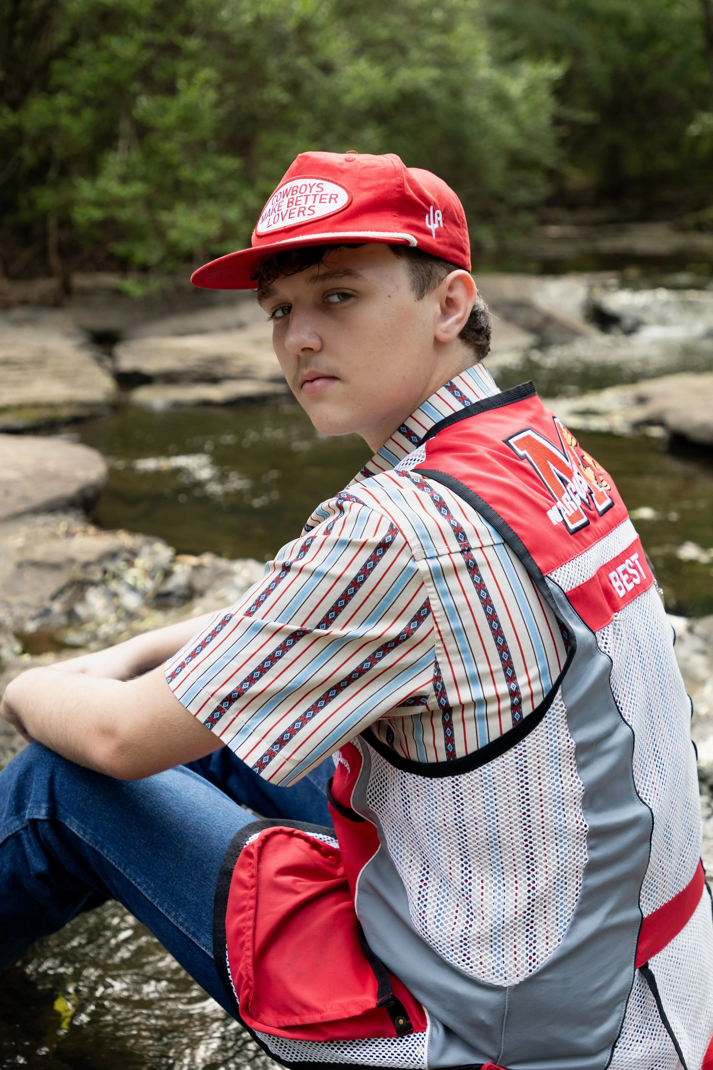 Teen boy in striped shirt and red cap sitting by a creek in a wooded area.