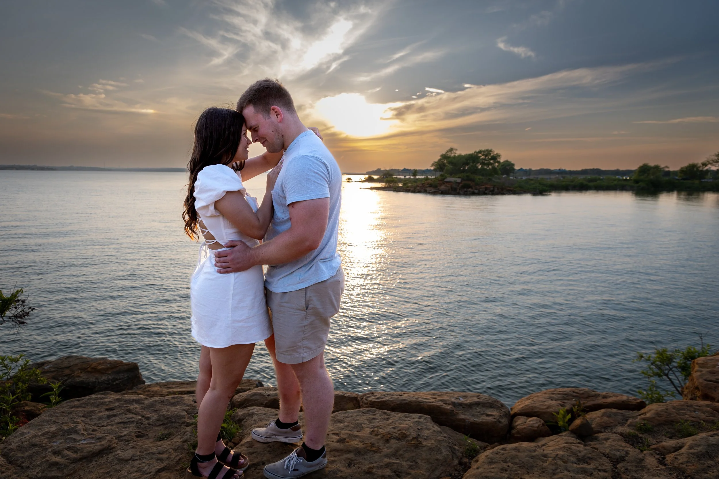 A couple stands close together on rocks by the water during sunset, touching foreheads and holding each other affectionately.