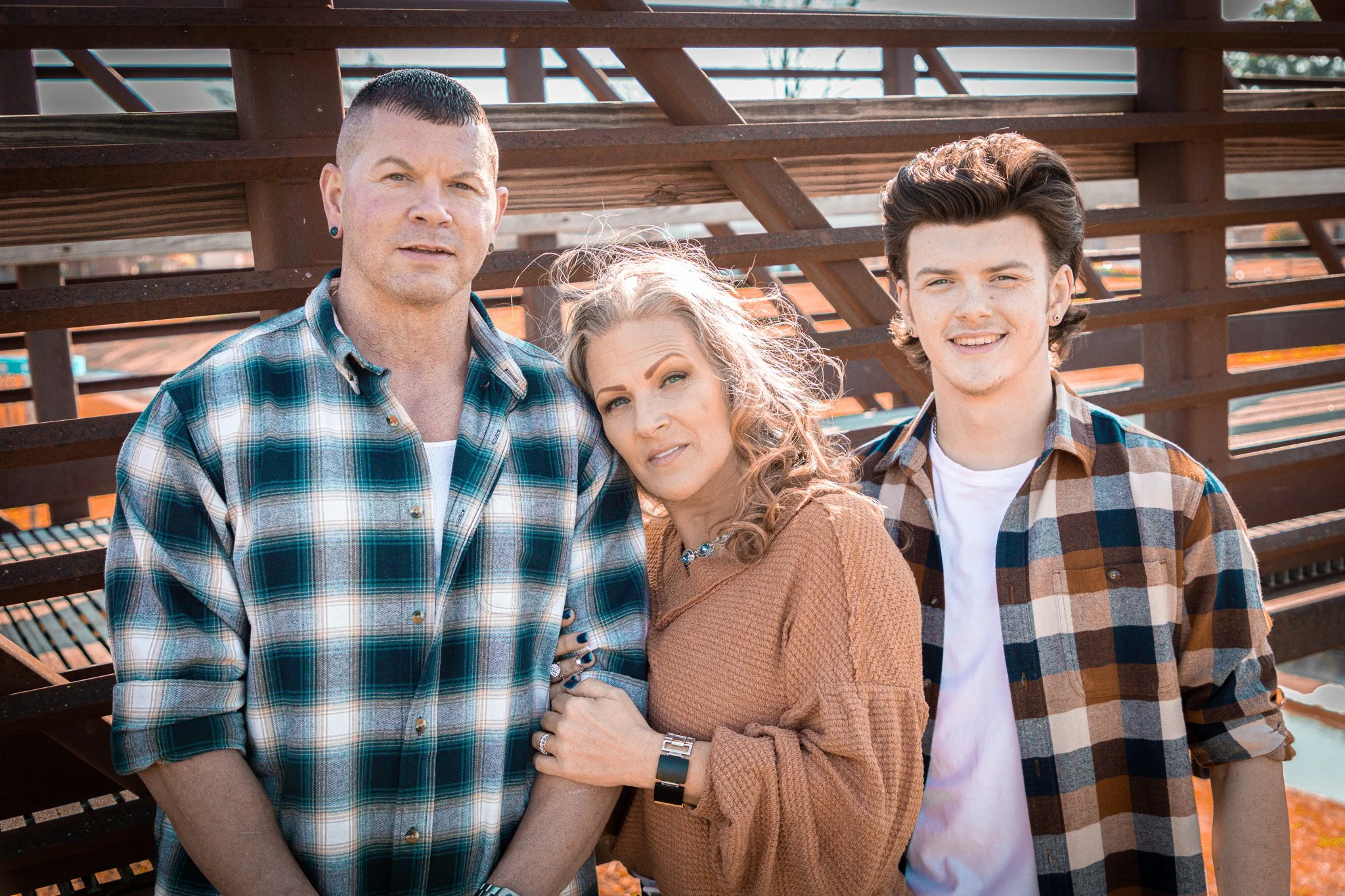 A family of three posing outdoors in front of metal bleachers, with the man on the left wearing a green plaid shirt, the woman in the middle wearing a brown sweater, and the young man on the right wearing a brown and blue plaid shirt.