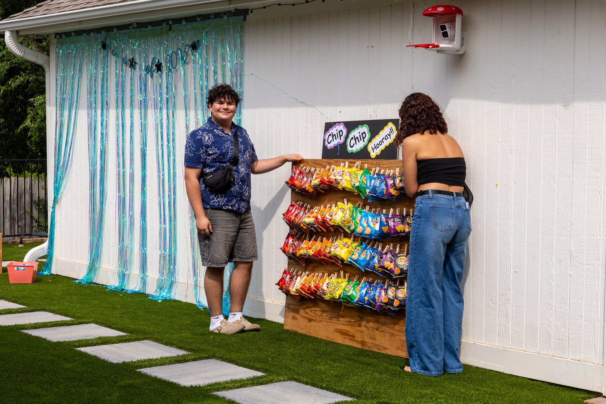 Two people at a snack booth with colorful chips on display, a sign with 'Chip Chip Hooray!!' above, in an outdoor setting with a decorative backdrop of blue streamers.