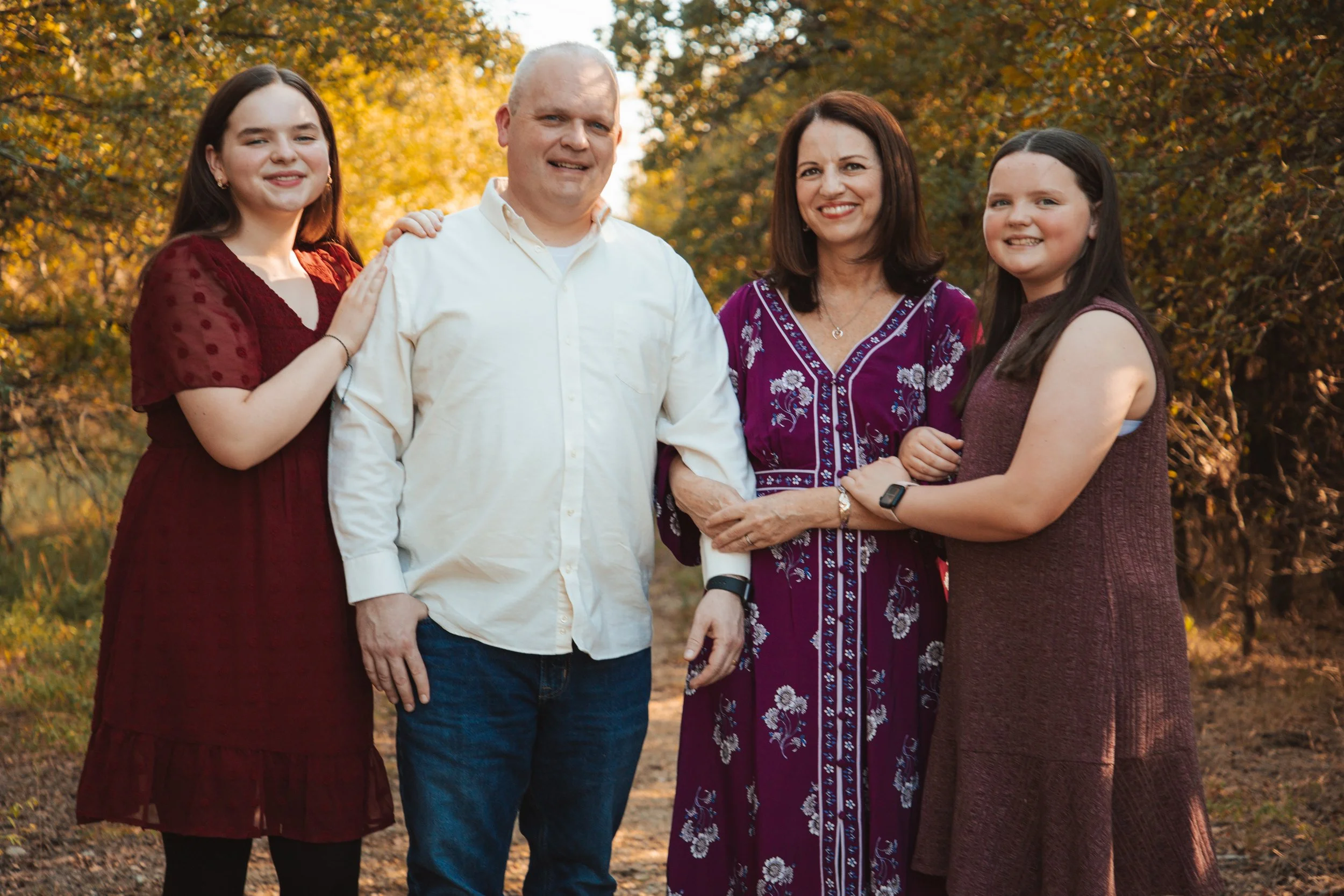 A family of four smiling outdoors in fall, with trees and yellow leaves in the background. The father and mother are in the center, with two daughters on either side.