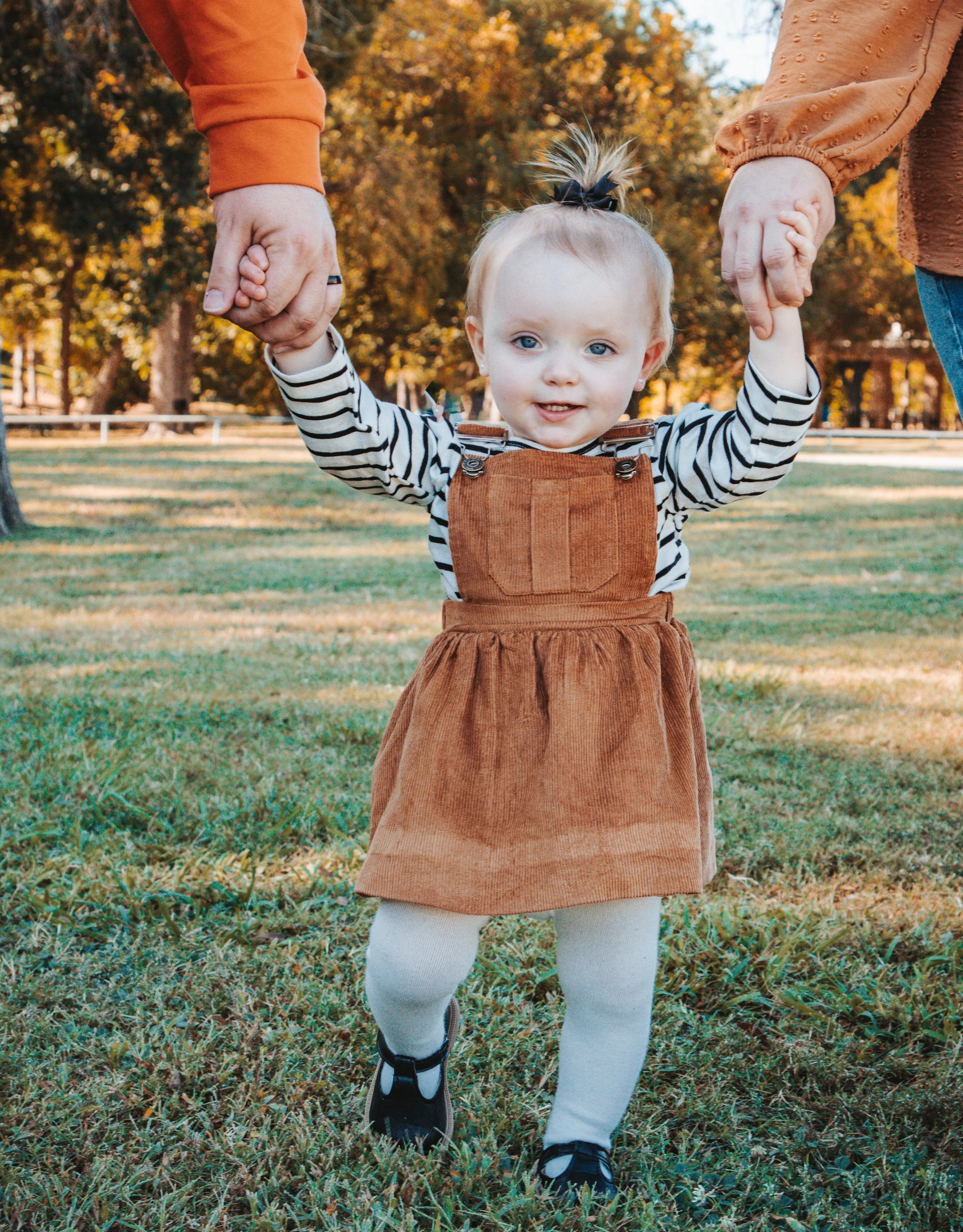 A young child with blond hair, blue eyes, wearing a striped shirt, brown corduroy dress, white tights, and black shoes, holding hands with two adults outdoors on a grassy field in autumn.