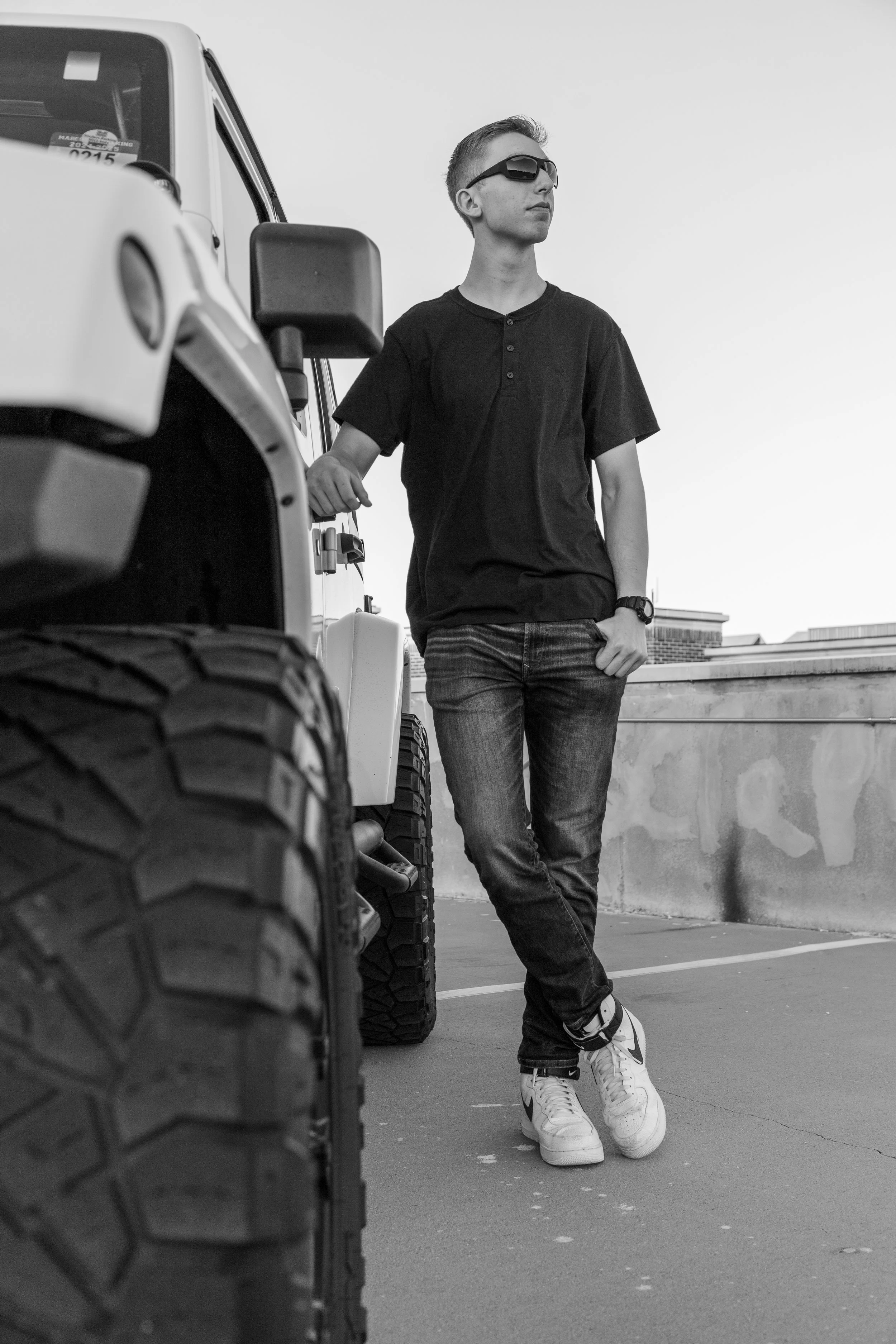 A young man wearing sunglasses, a black t-shirt, jeans, and sneakers leaning against a large off-road vehicle on a rooftop parking lot in black and white.