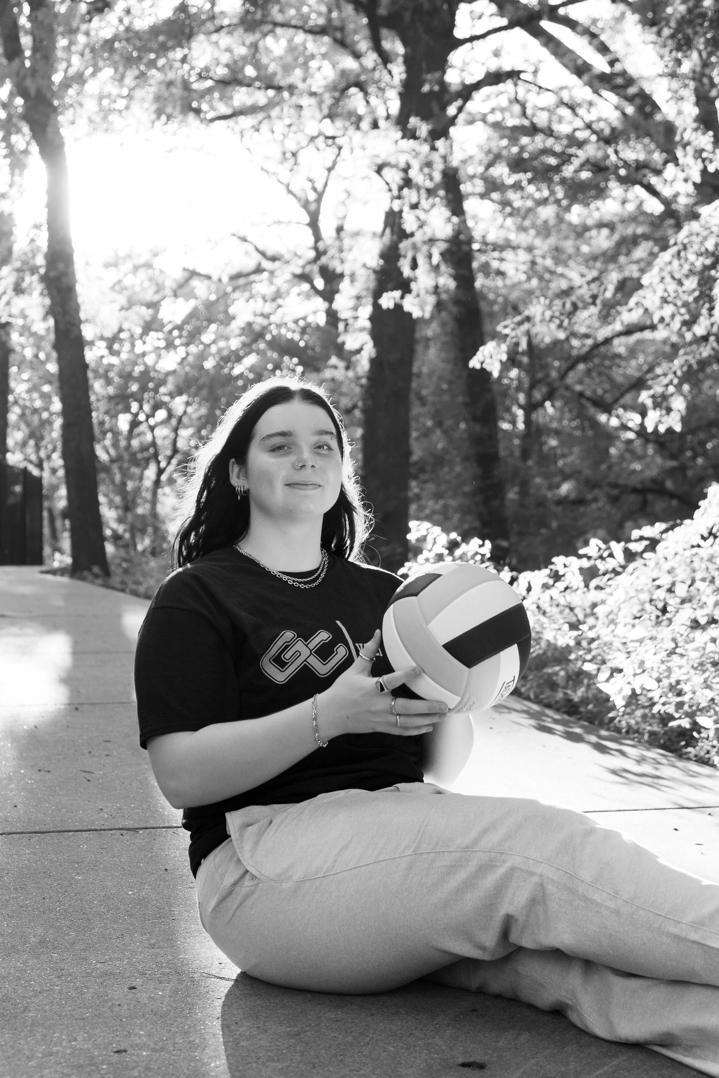 A young woman with dark hair, wearing a black t-shirt and light-colored pants, sitting on the sidewalk outdoors. She is holding a volleyball and is surrounded by trees with sunlight filtering through the branches.