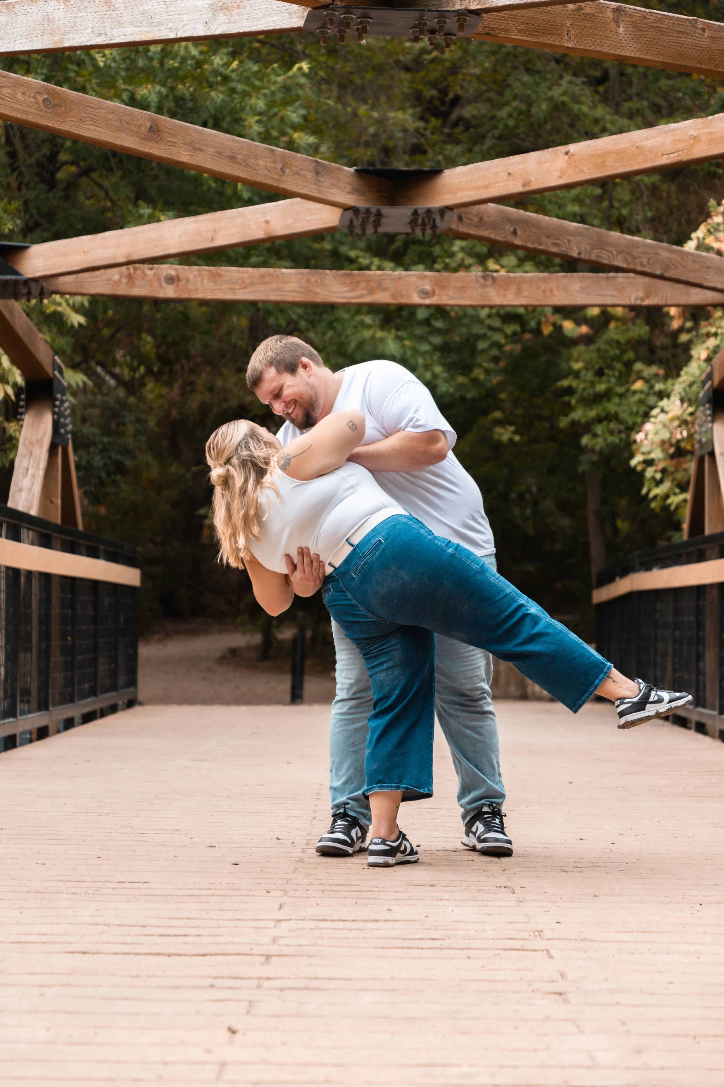 A man and woman dancing and smiling on a wooden bridge surrounded by nature.