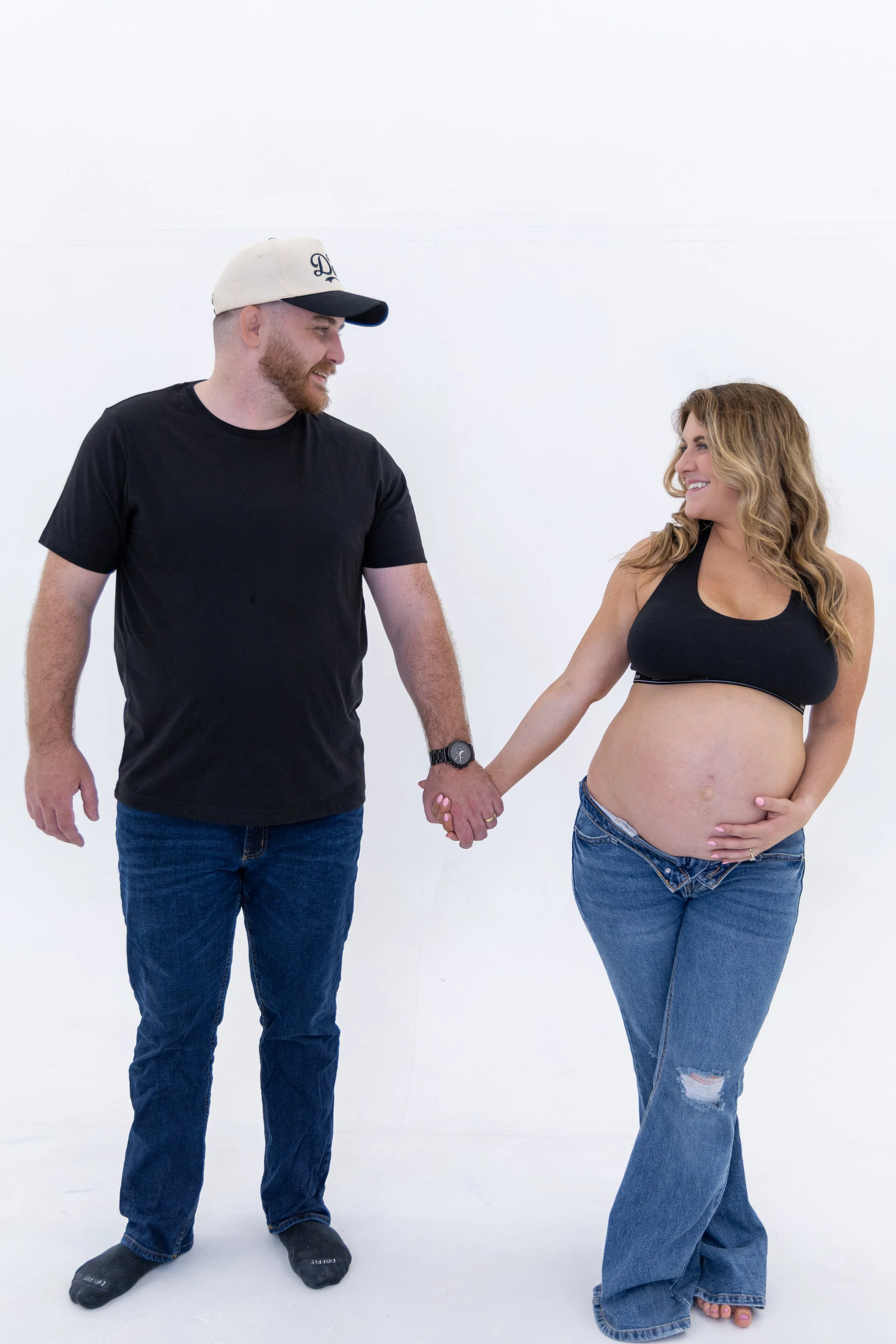 A man and a pregnant woman holding hands, smiling at each other against a plain white background.