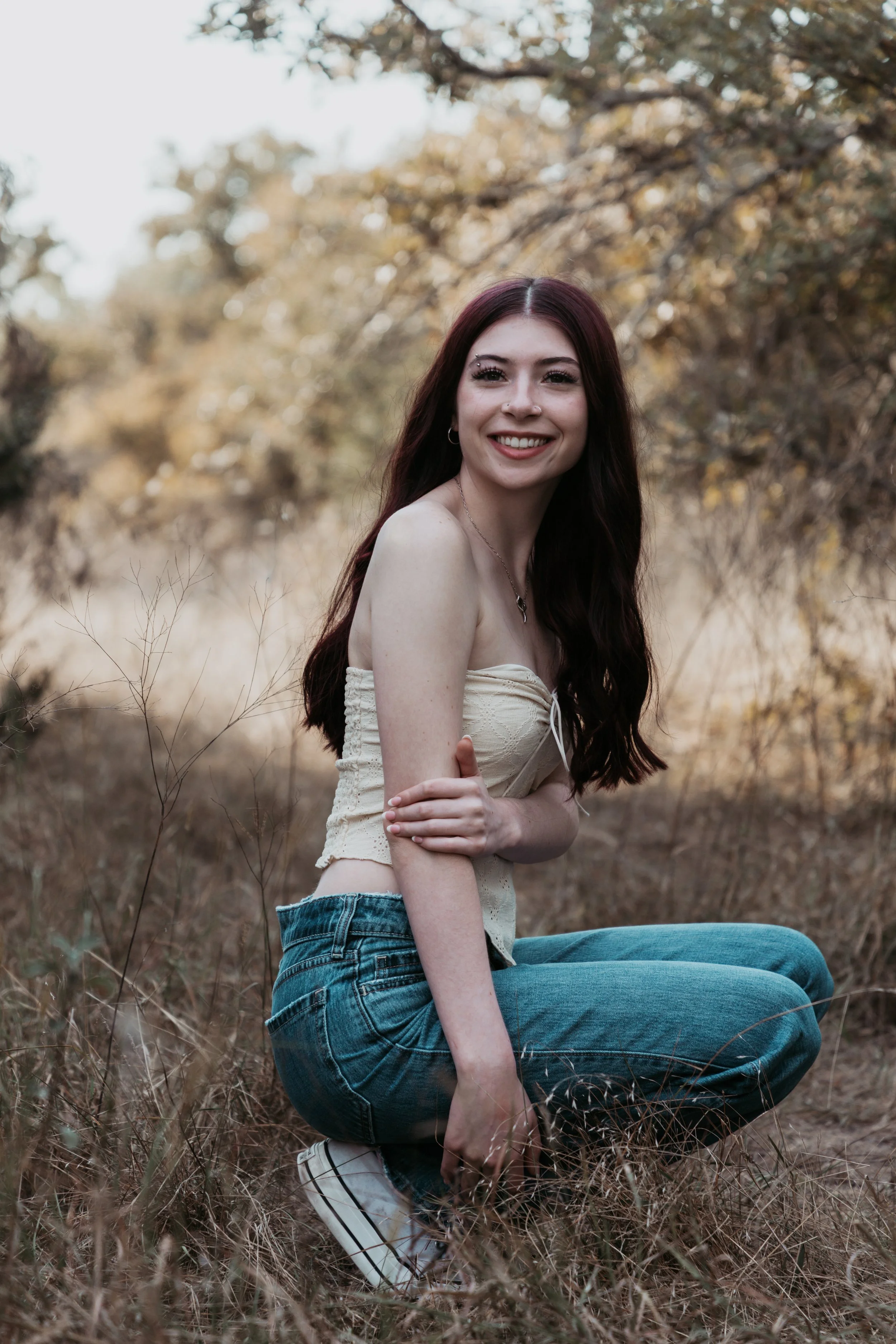 A young woman with long reddish-brown hair, smiling, sitting on the ground in a field with tall grass and trees in the background, wearing a strapless beige top, blue jeans, and sneakers.
