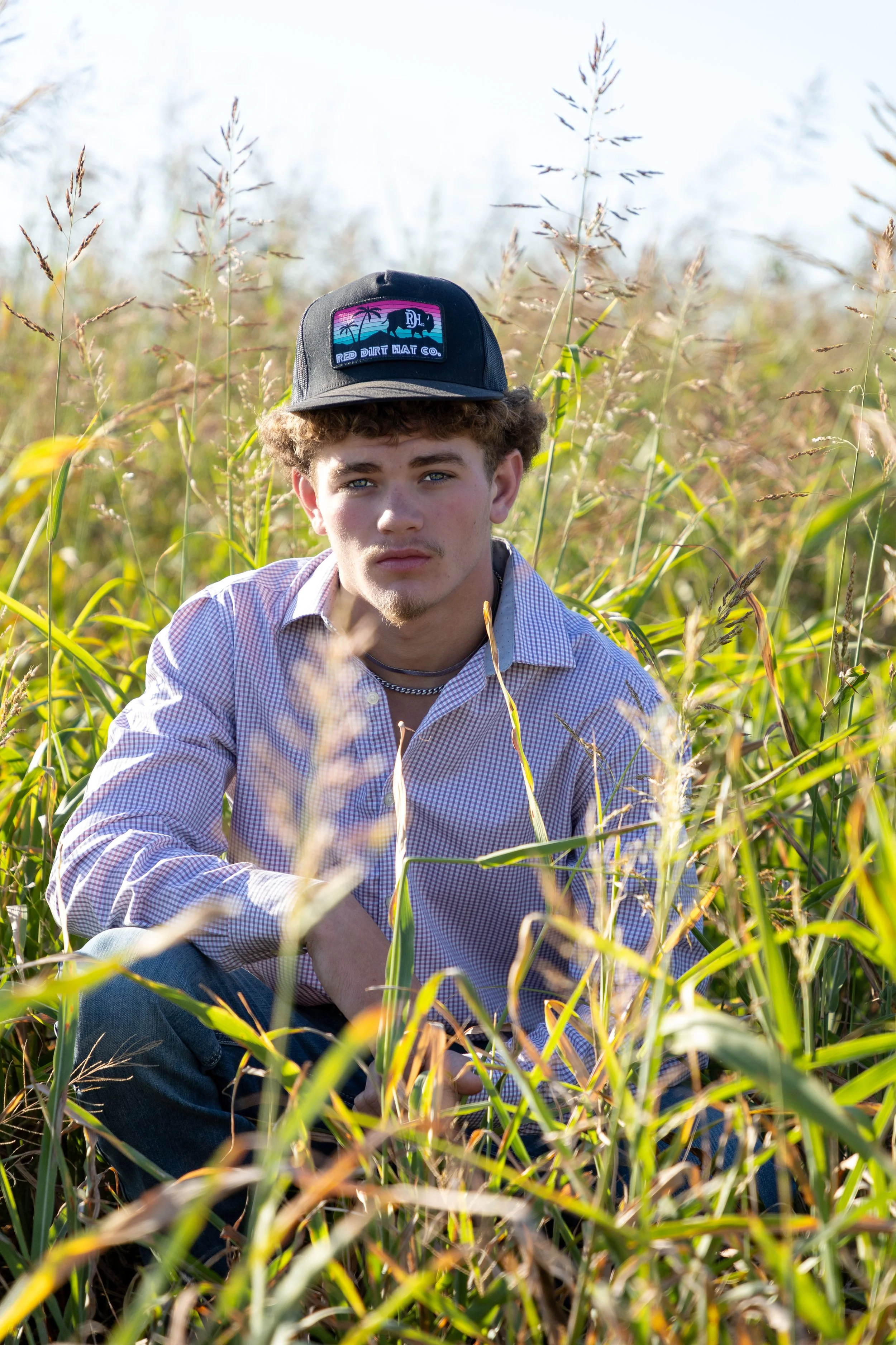 Young man sitting in a field of tall grass, wearing a checkered shirt, a black cap with a tropical patch, and earrings, looking directly at the camera.