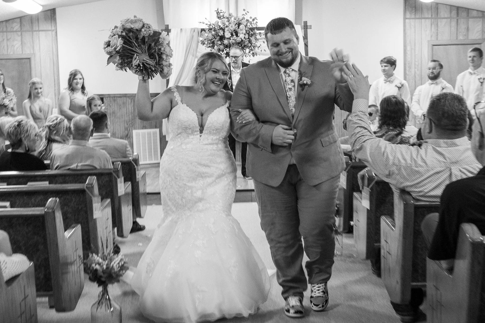 A bride and groom smiling and giving high-fives during their wedding ceremony inside a church, with guests seated and standing around them.