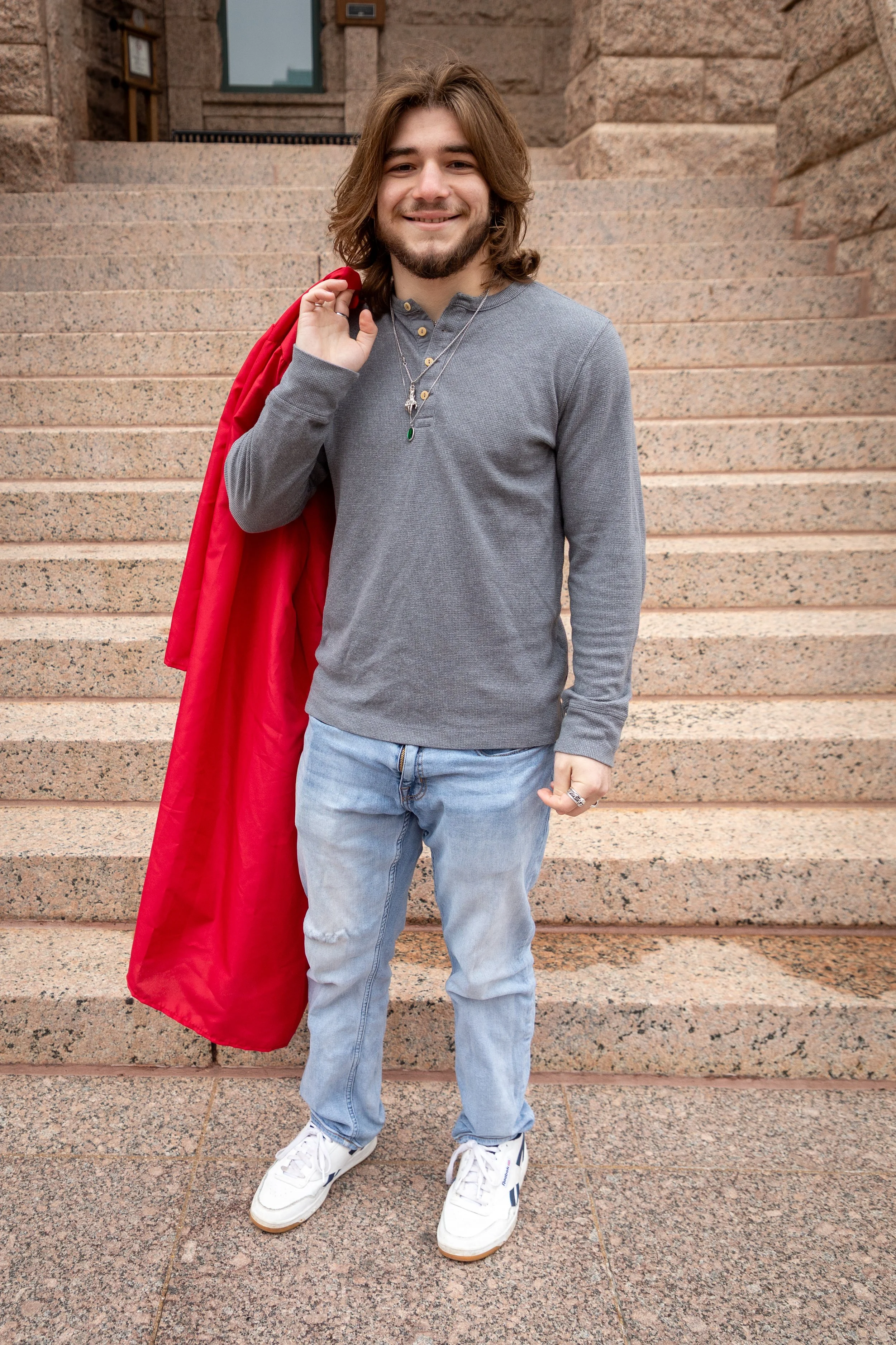 A young man with long hair stands on a brick staircase, smiling, holding a red jacket over his shoulder, wearing a gray long-sleeve shirt, light blue jeans, and white sneakers.