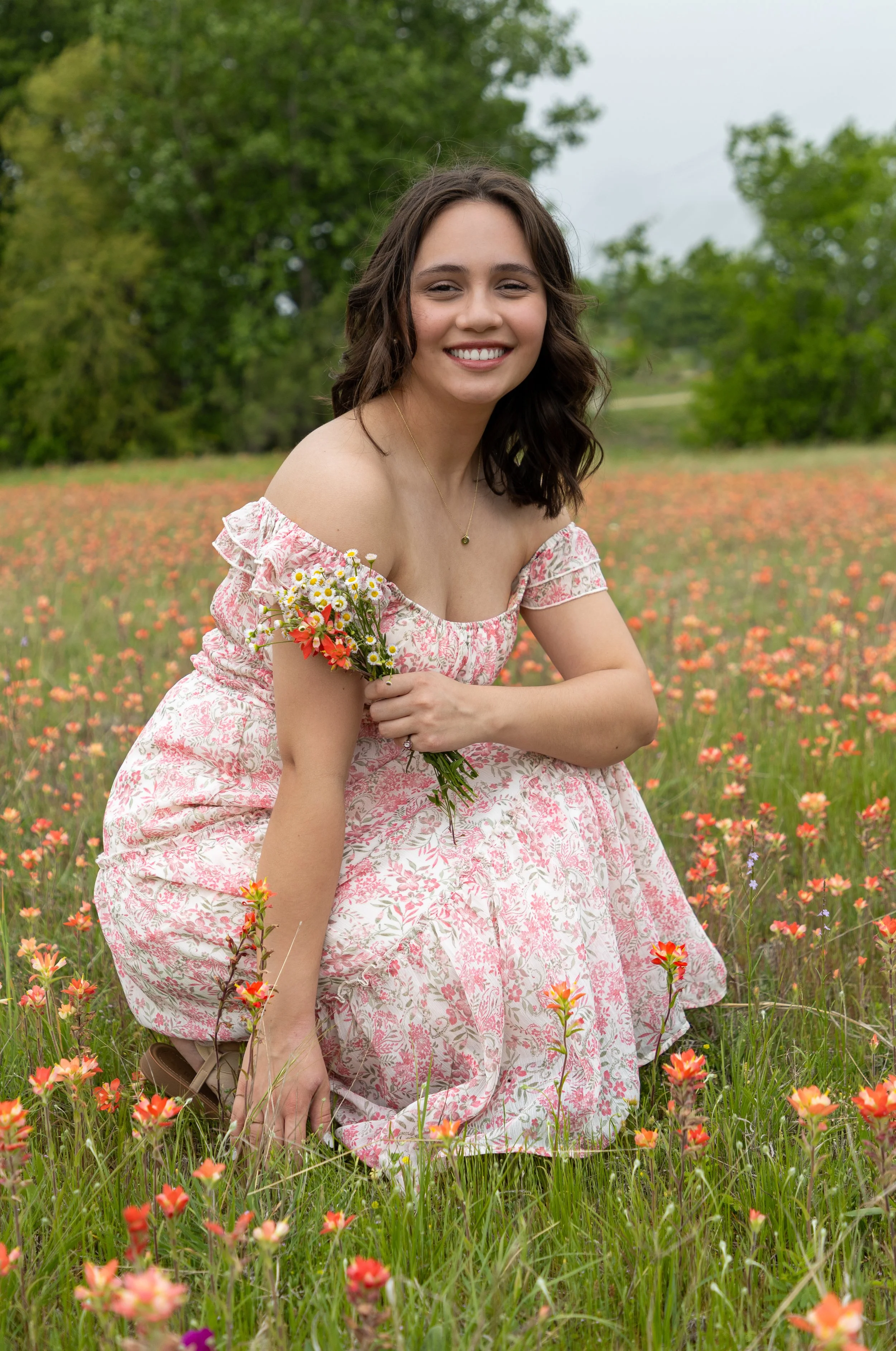 Young woman in a pink floral dress kneeling in a field of blooming flowers, holding a small bouquet, smiling, with green trees and an overcast sky in the background.