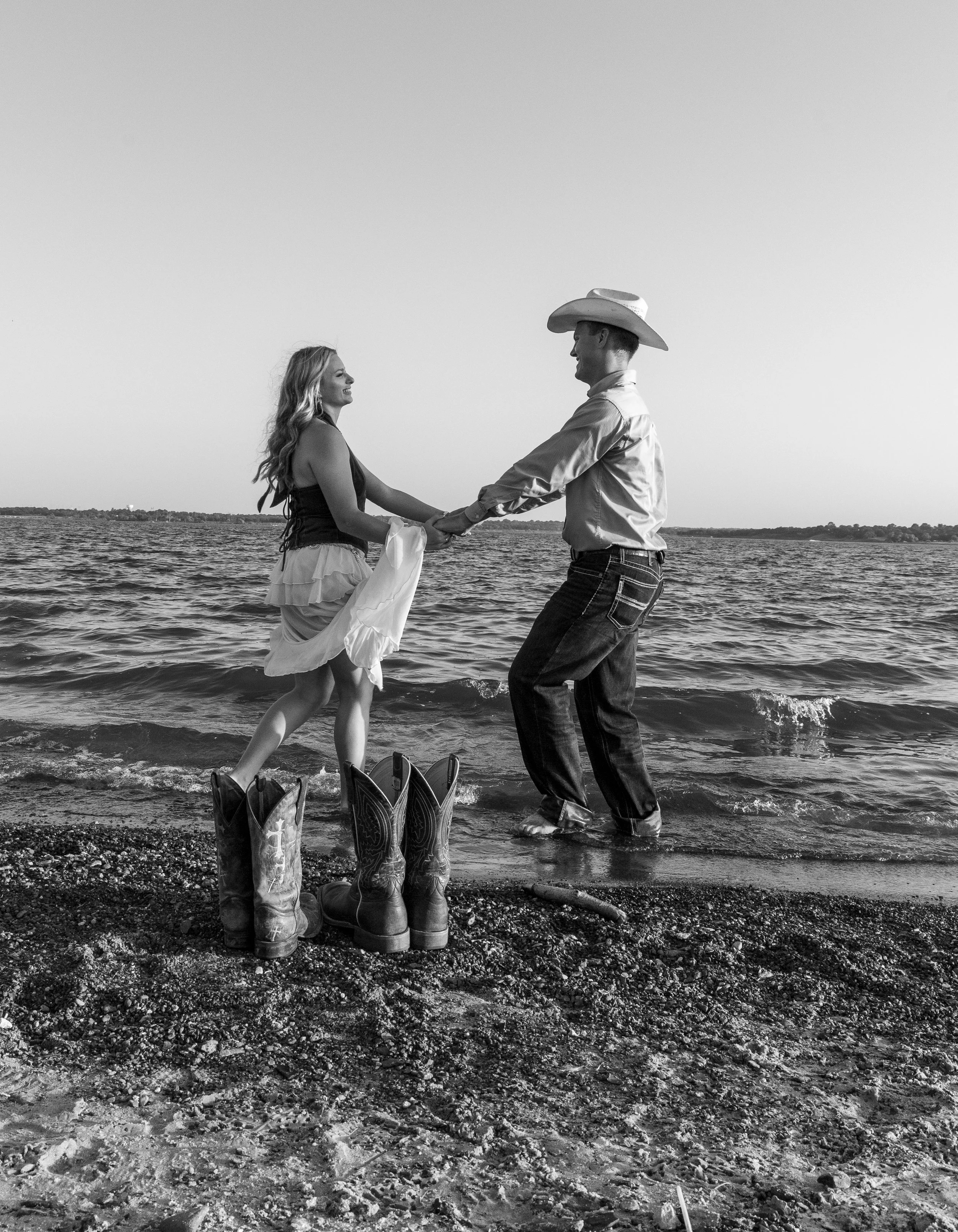 A black and white photo of a man and woman holding hands and dancing by the water. The woman is wearing cowboy boots and a dress, while the man is wearing a cowboy hat, shirt, and jeans. There are additional cowboy boots on the ground and the scene i