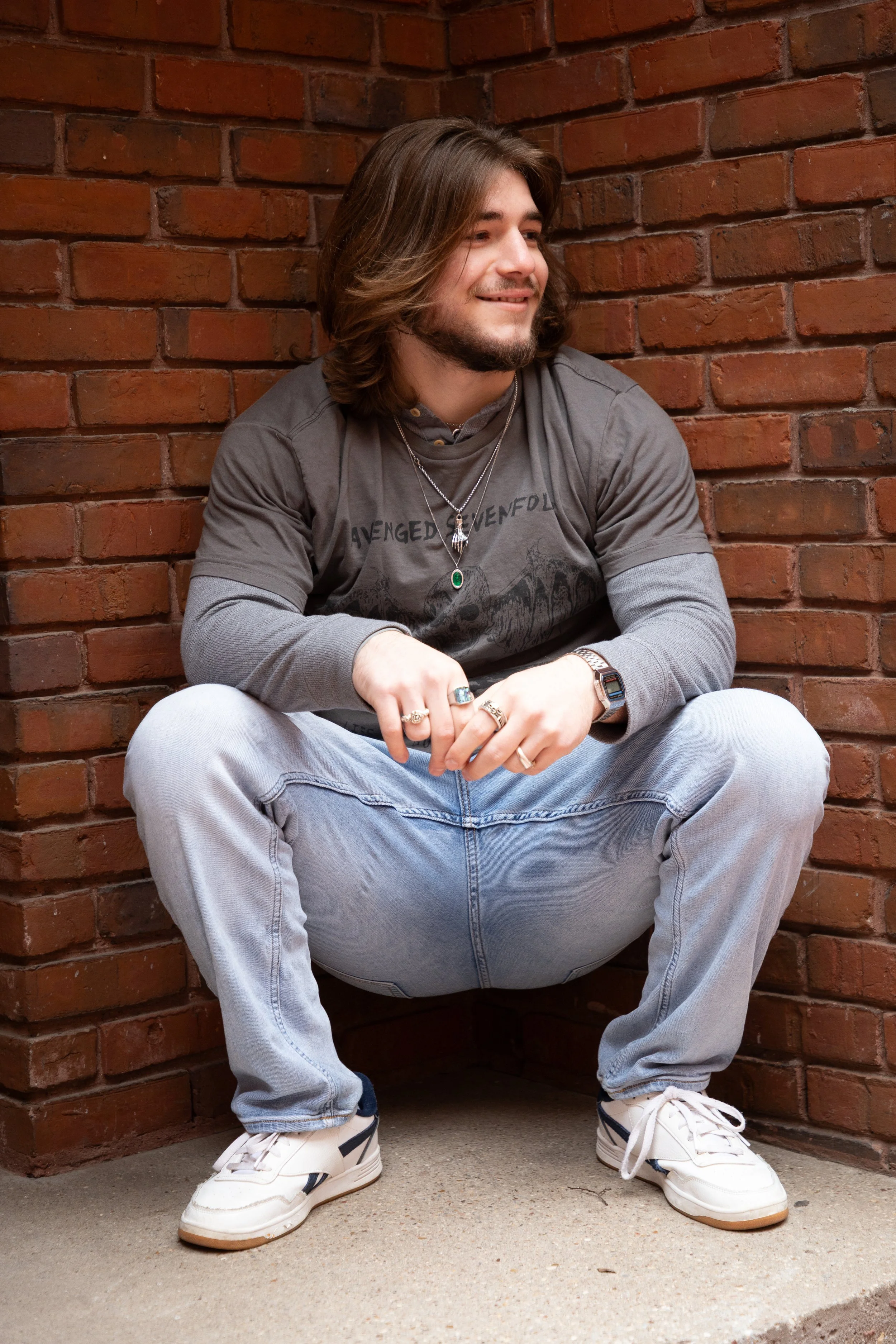 A young man with shoulder-length brown hair and a beard sitting on the ground against a brick wall, smiling and looking to the side.