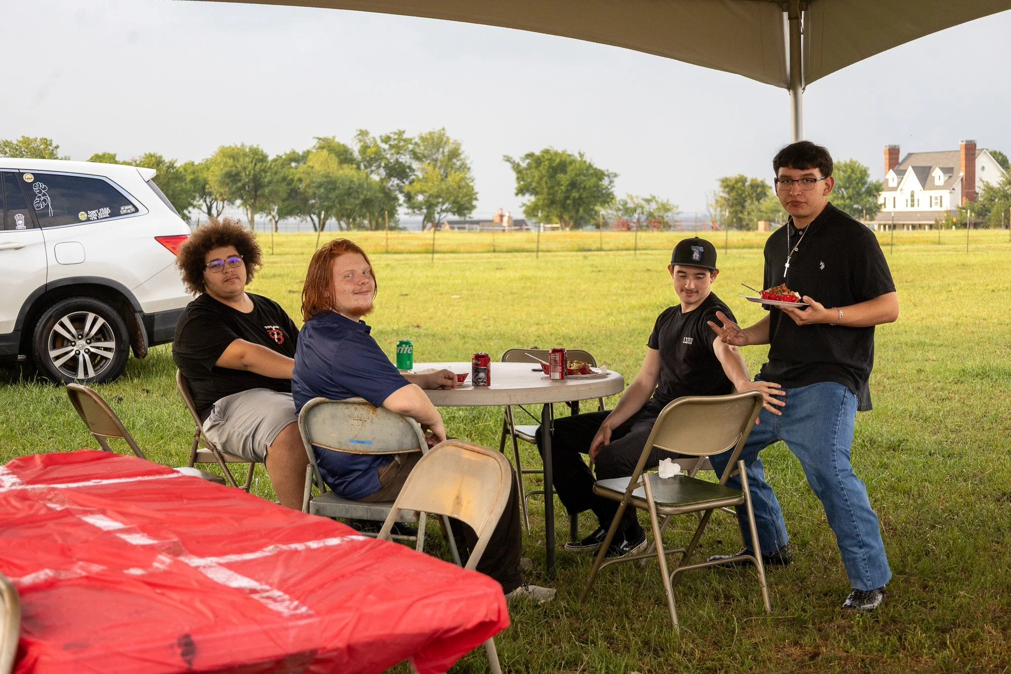 Four young men sitting around a white picnic table under a beige canopy outdoors. One young man is standing, holding food and making a peace sign. The other three are sitting, two facing the camera and one with his back turned. There are soda cans an