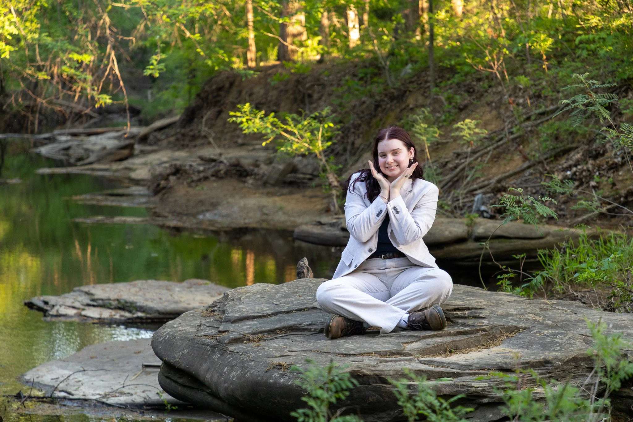 A woman sitting cross-legged on a large rock in a forested area near a creek, smiling with her hands on her cheeks, wearing a white suit.