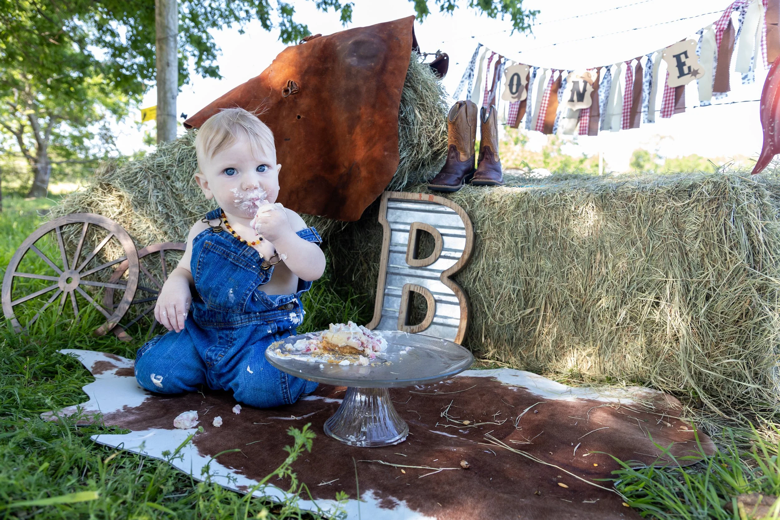 A young child with blond hair and blue eyes sitting outdoors on a animal print blanket, eating cake with white frosting. The child is wearing denim overalls and a beaded necklace. Behind the child is a rustic farm-themed background with hay, a decora