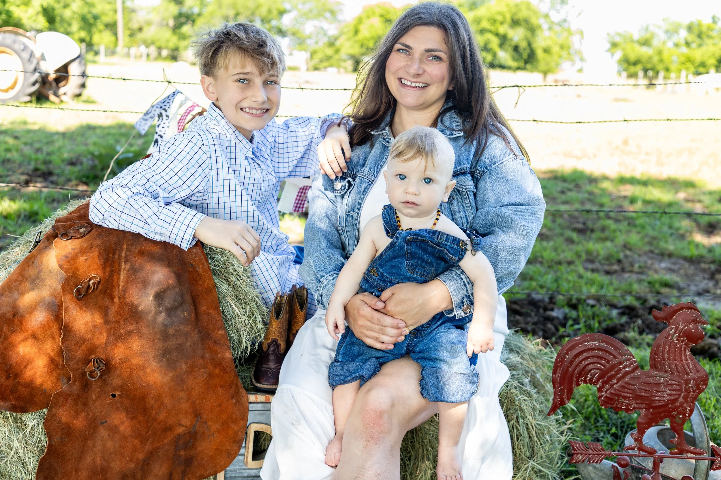 A woman with dark hair smiling outside, sitting on hay bales, with two young children. One child, a boy with light hair, is leaning on her and smiling at the camera. The other, a toddler with light hair, is sitting on her lap, wearing denim overalls,