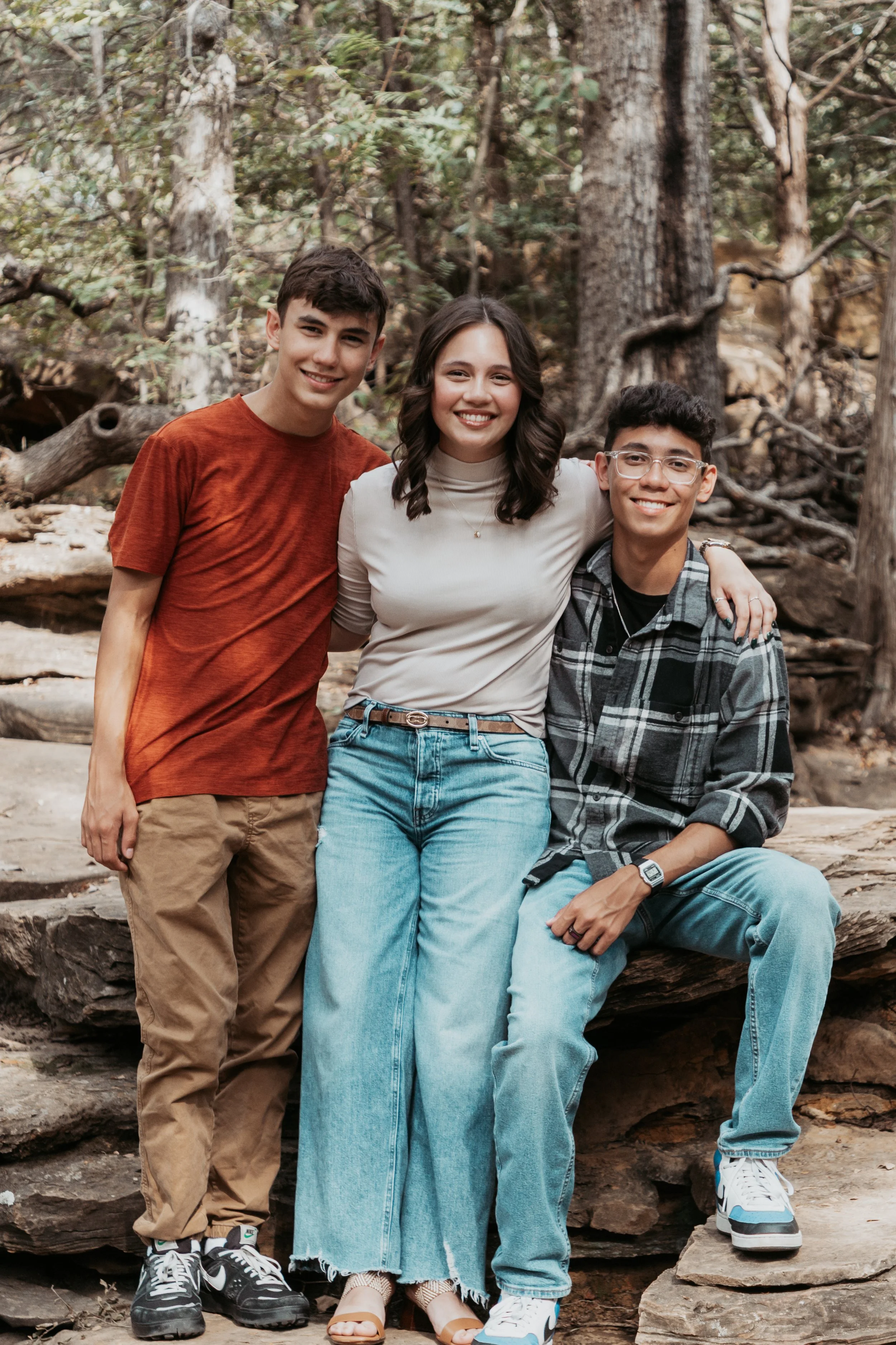 Three teenagers in casual clothing standing outdoors in a wooded area, smiling for the camera.