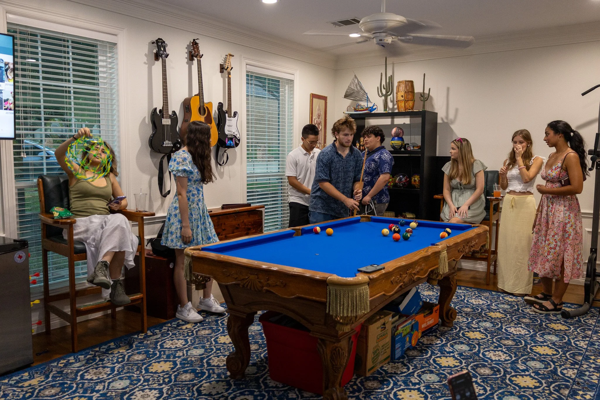 A group of people gathered around a pool table in a living room, with guitars hanging on the wall, and various decorations, some holding drinks and socializing.