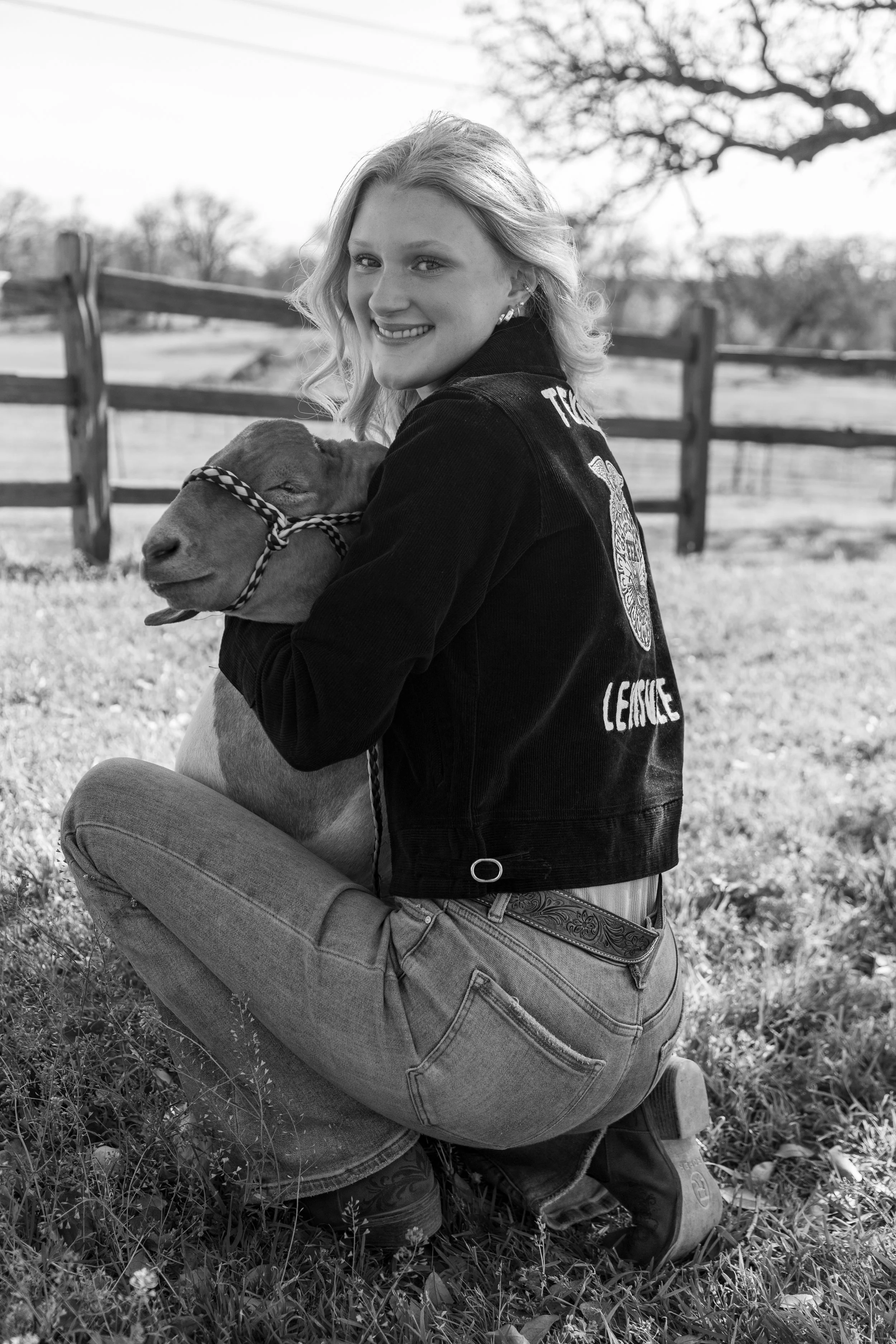 A smiling woman in jeans and a jacket kneels on grass, hugging a lamb outdoors with a wooden fence and trees in the background.