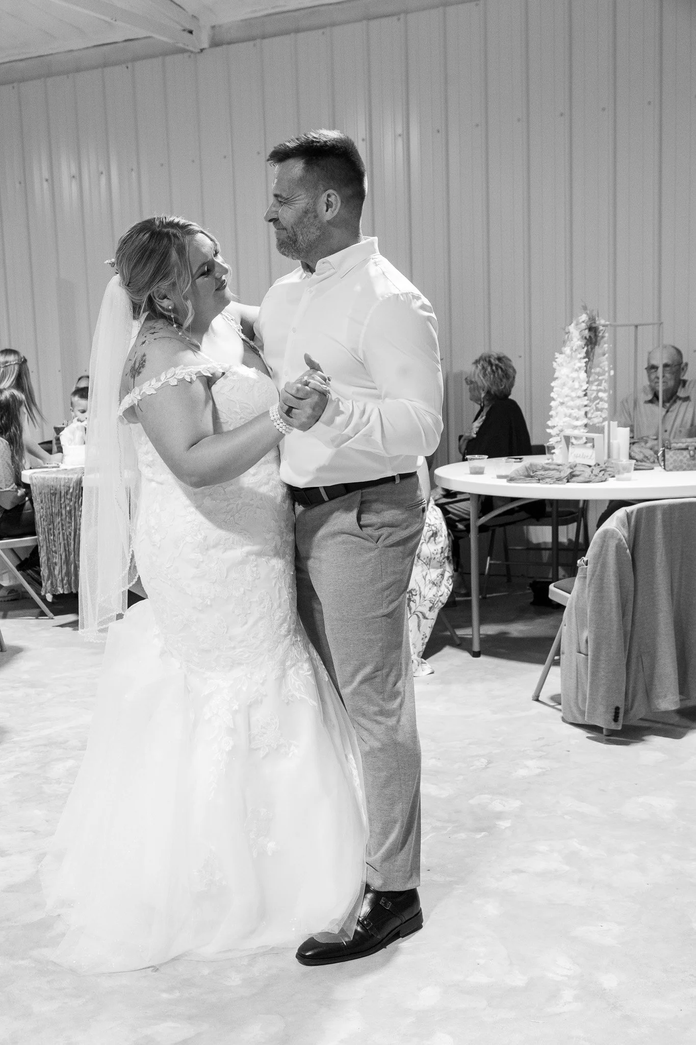 A couple sharing their first dance at a wedding reception in a rustic venue with wooden walls and guests seated at tables in the background.