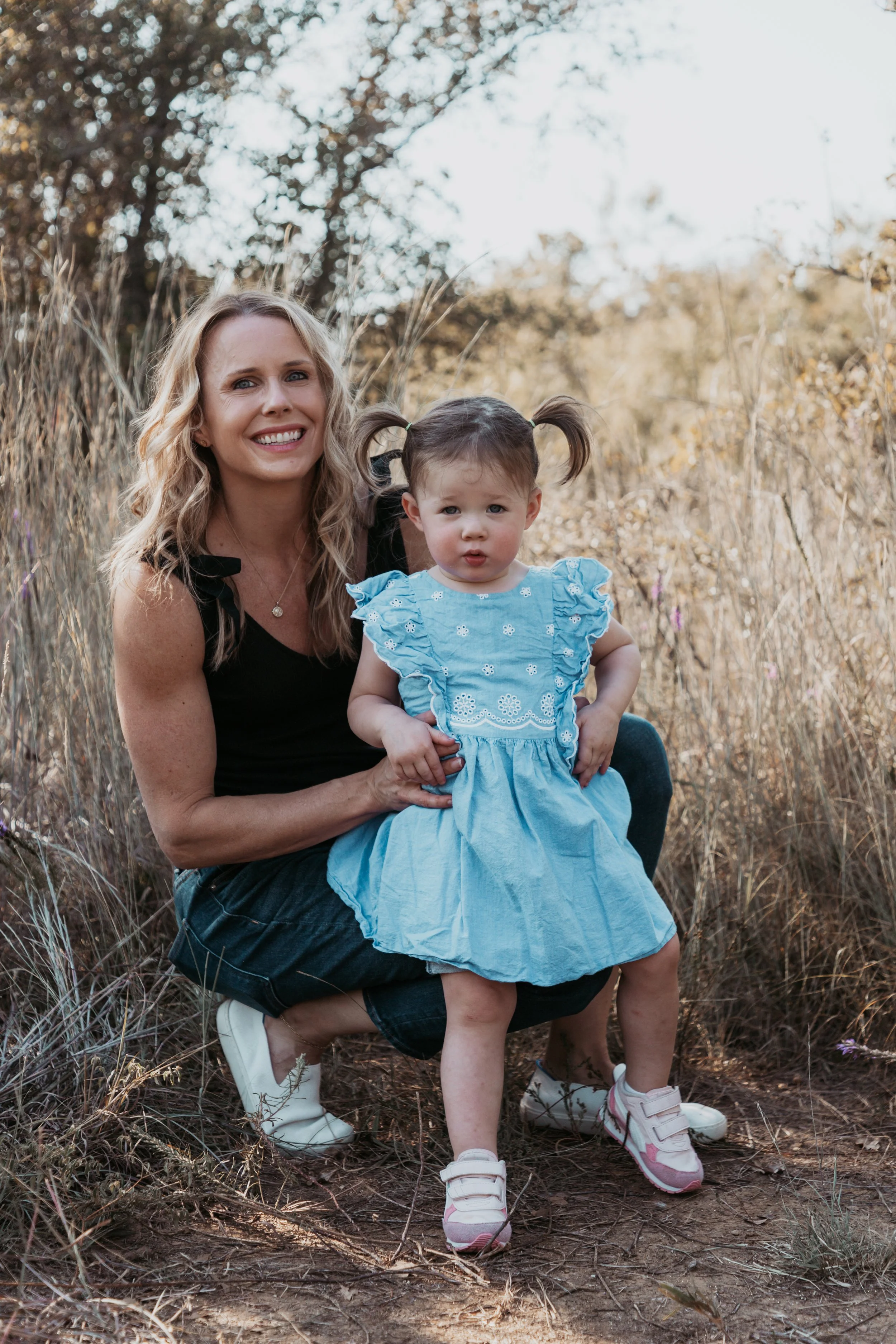 A woman with blonde hair smiling and a young girl with pigtails and a surprised expression, outdoors in a dry grassy area with trees in the background.