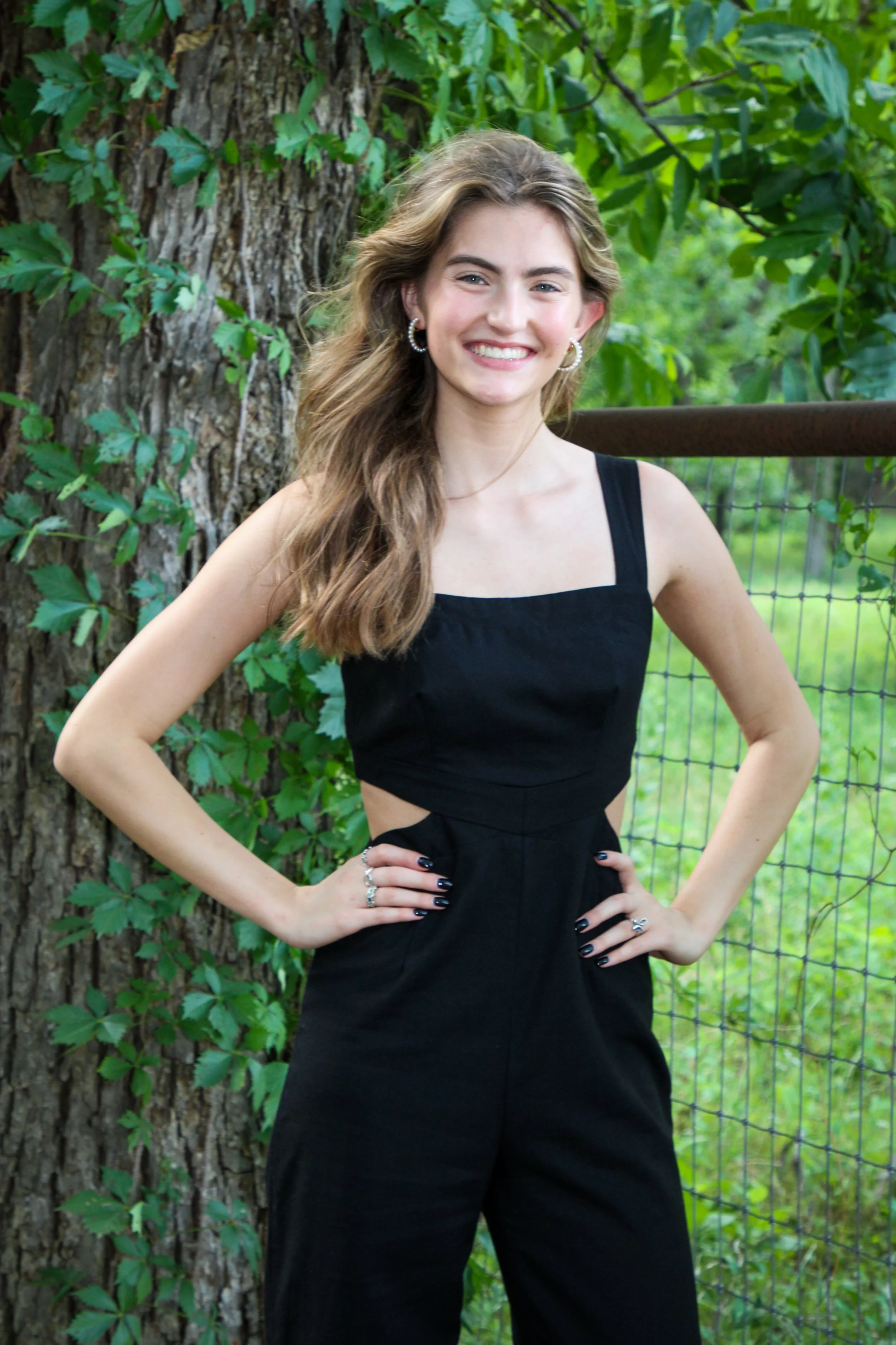 A young woman with long wavy hair wearing a black sleeveless jumpsuit standing outdoors next to a large tree with green leaves, hands on her hips, smiling.