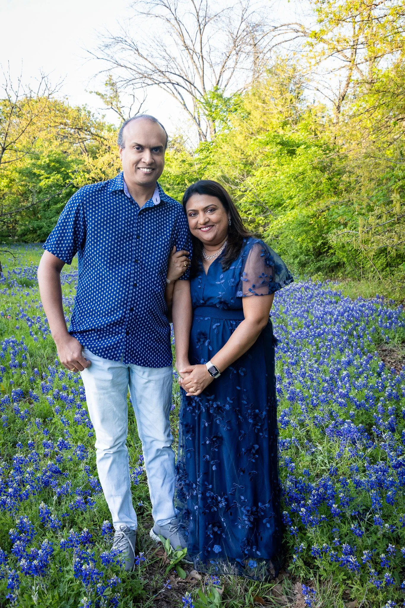 A man and woman standing close together in a field of blue flowers with green trees and clear sky in the background.