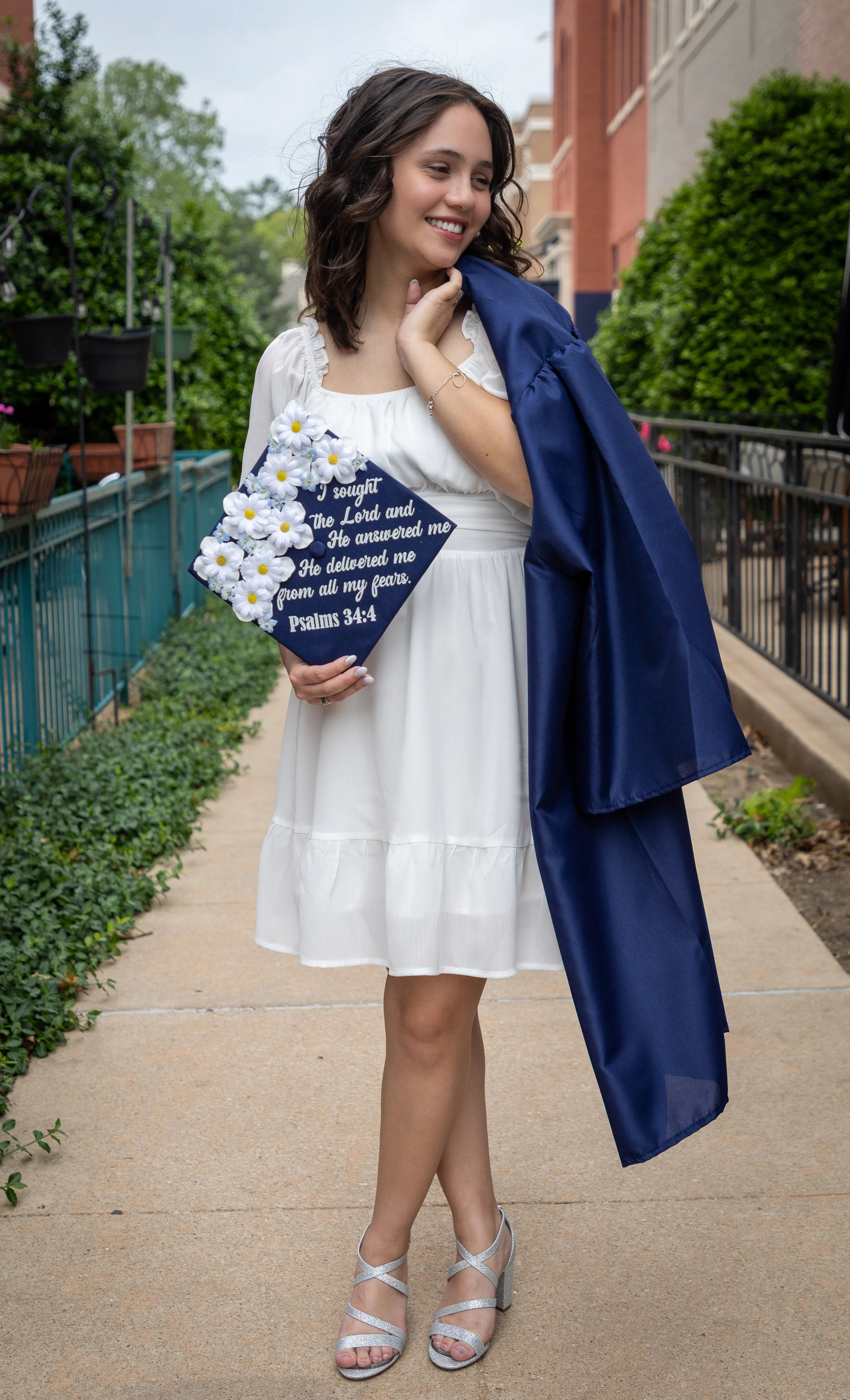 A young woman in a white dress and silver heels holding a graduation cap with a Psalms 34:4 scripture, standing outdoors on a sidewalk with greenery and buildings in the background.