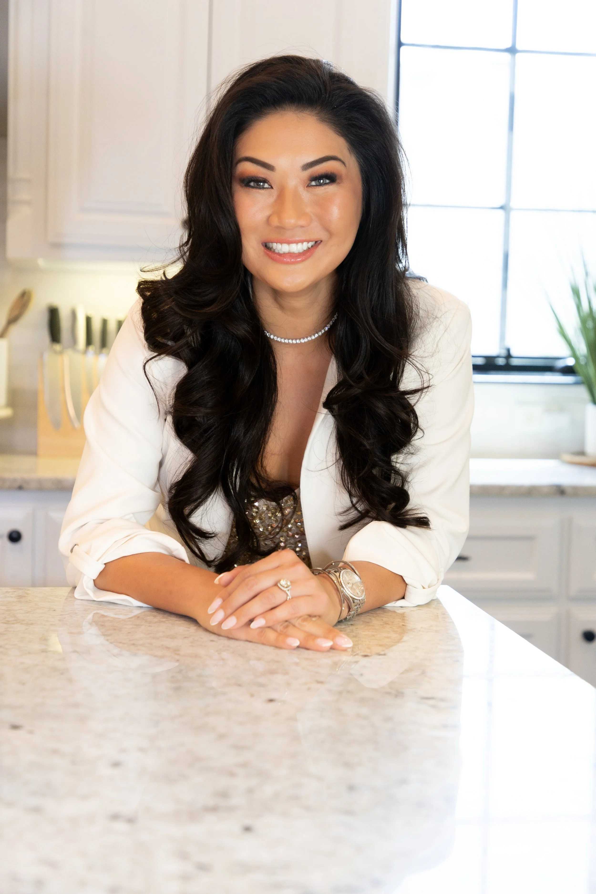 A smiling woman with long dark hair sitting at a kitchen counter, wearing a white blazer, jewelry, and a sequined top.