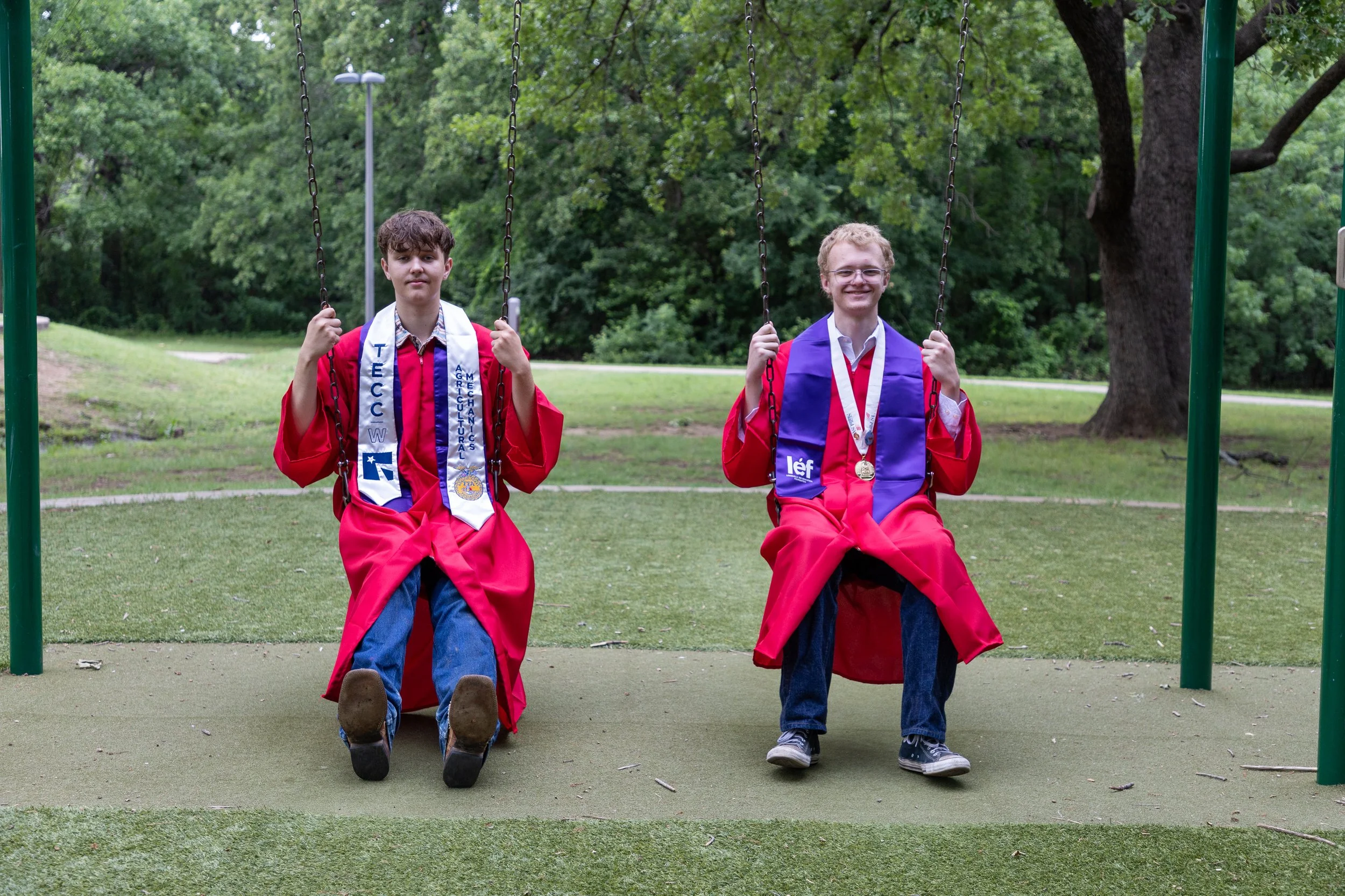 Two young men in red graduation gowns and stoles sitting on swings in a park, smiling with medals around their necks.